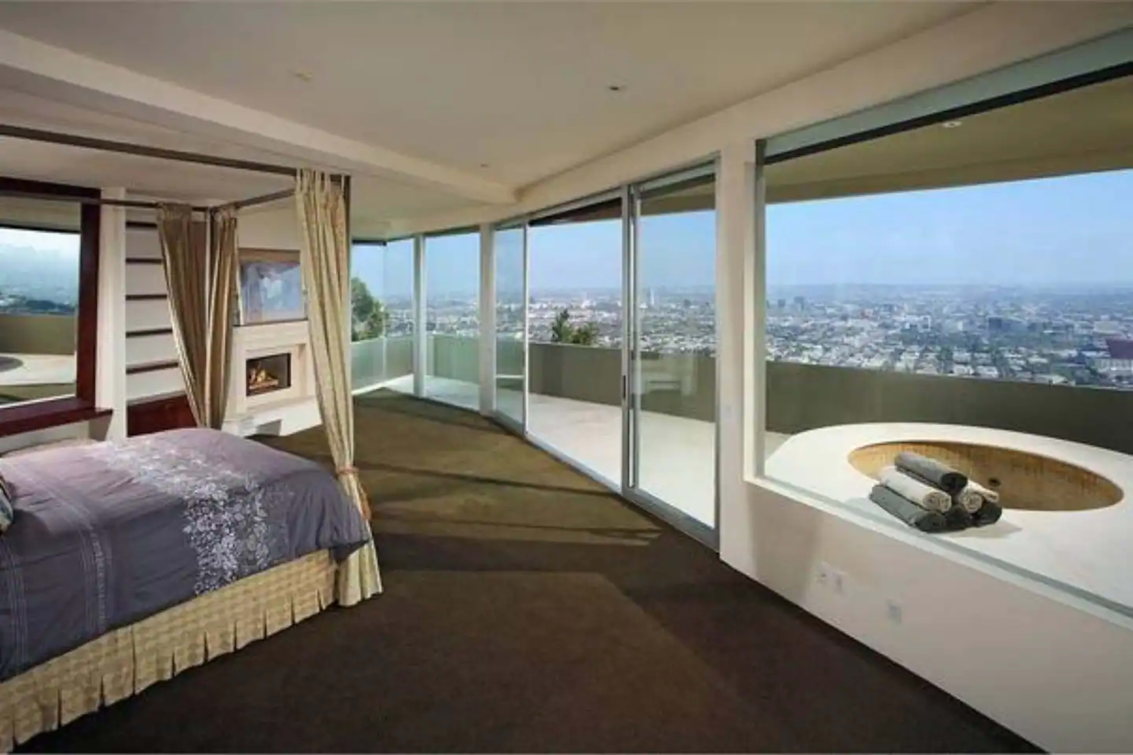 The image shows a modern bedroom with a large window overlooking a city view and an outdoor hot tub.
