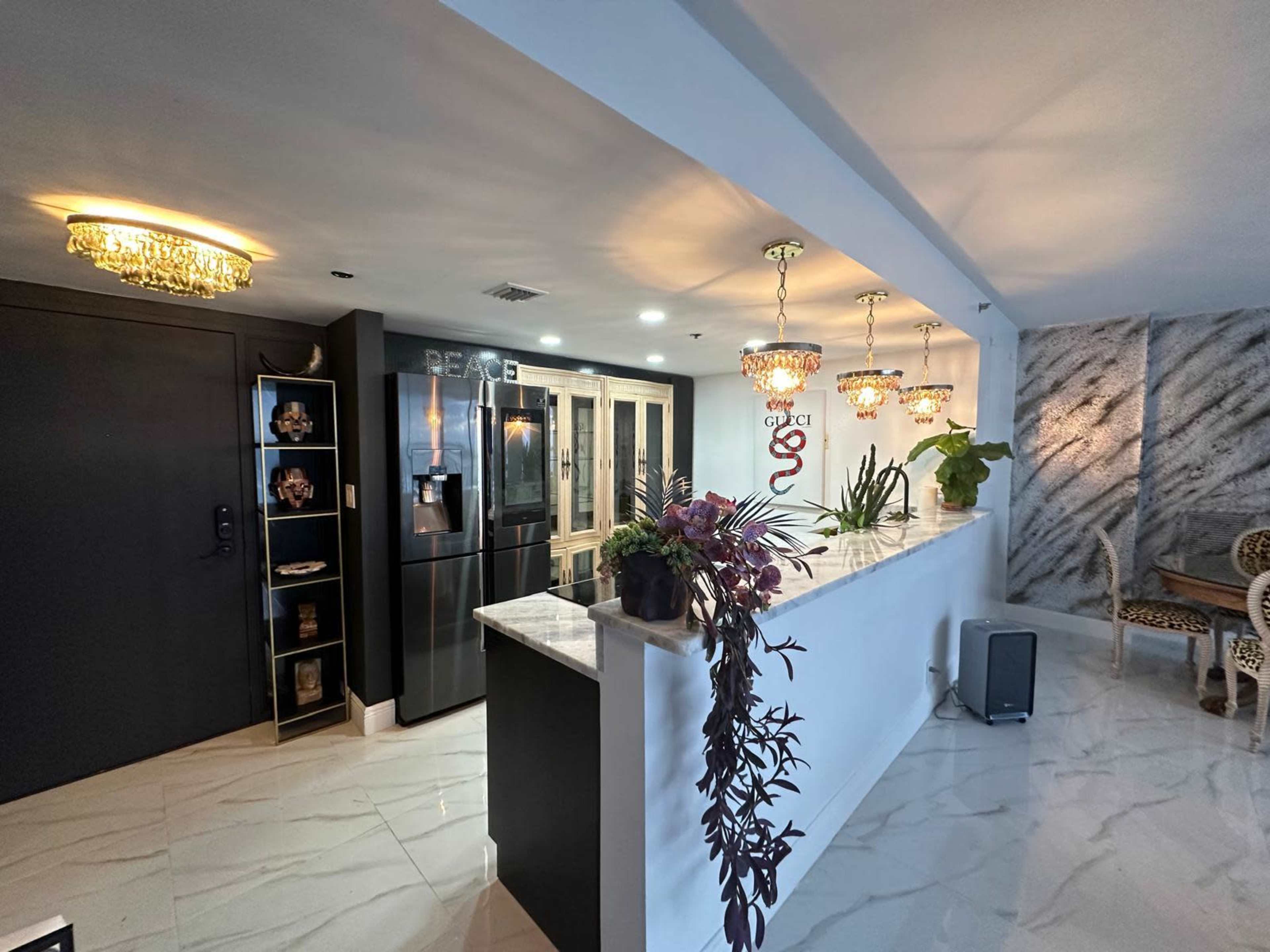 The image shows a modern kitchen with a black refrigerator, white countertops, and pendant lights, alongside decorative plants and ornate furniture in the dining area.