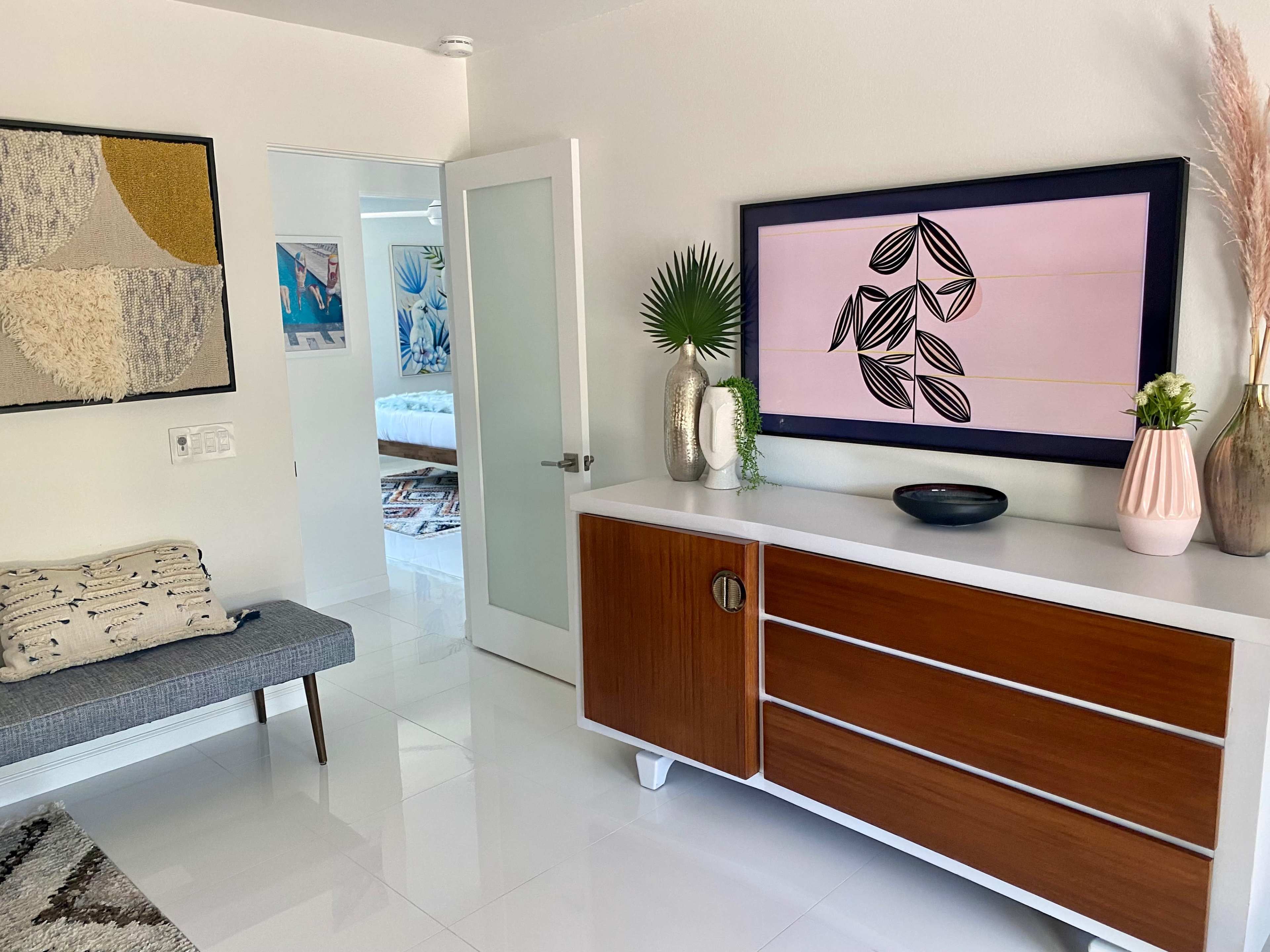 The image shows a modern entryway with a console table, artwork on the wall, and a seating area beside a door leading to another room.