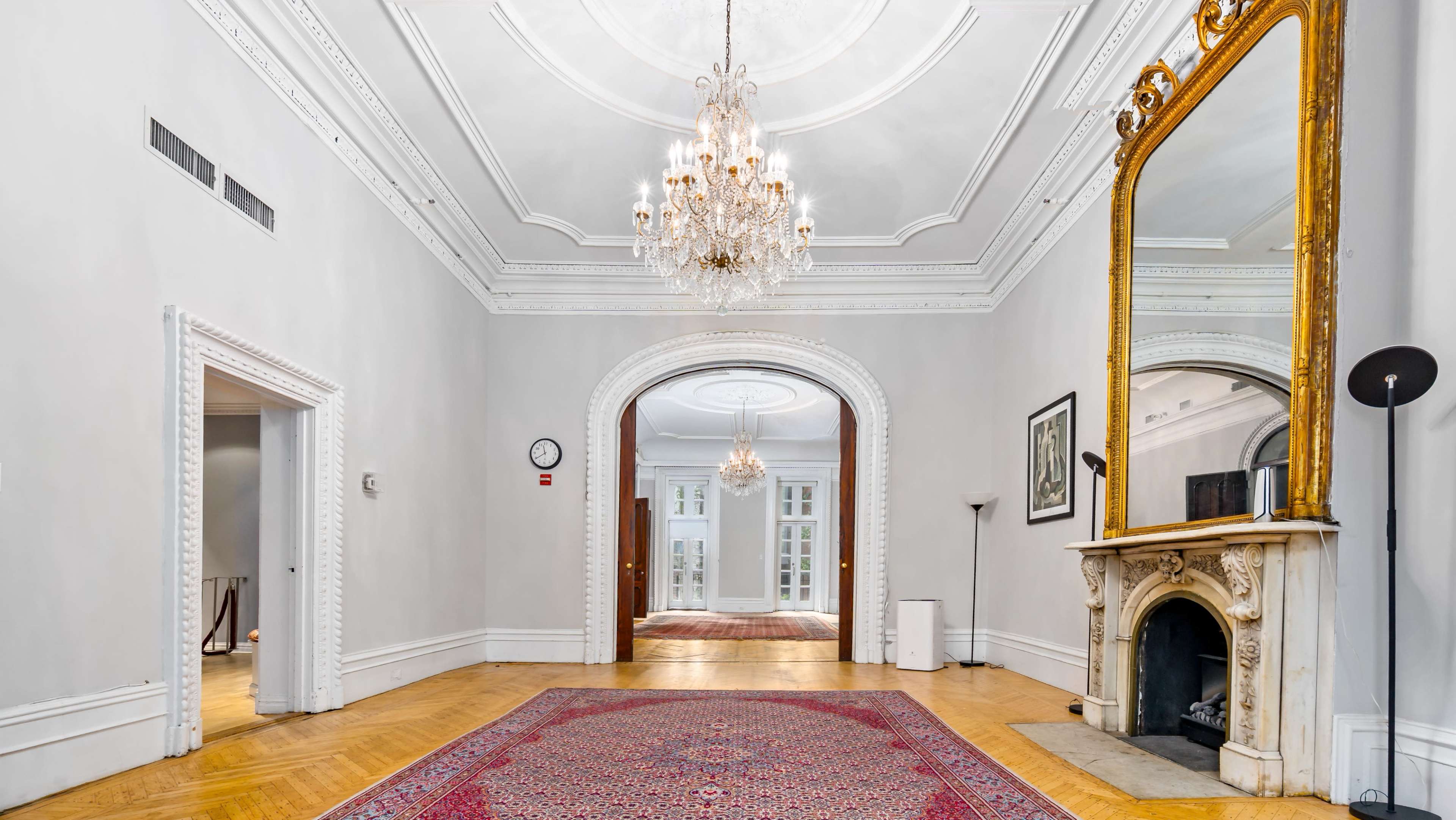 The image shows a spacious, elegantly designed hallway with a chandelier, ornate ceiling details, a large mirror, and a marble fireplace.