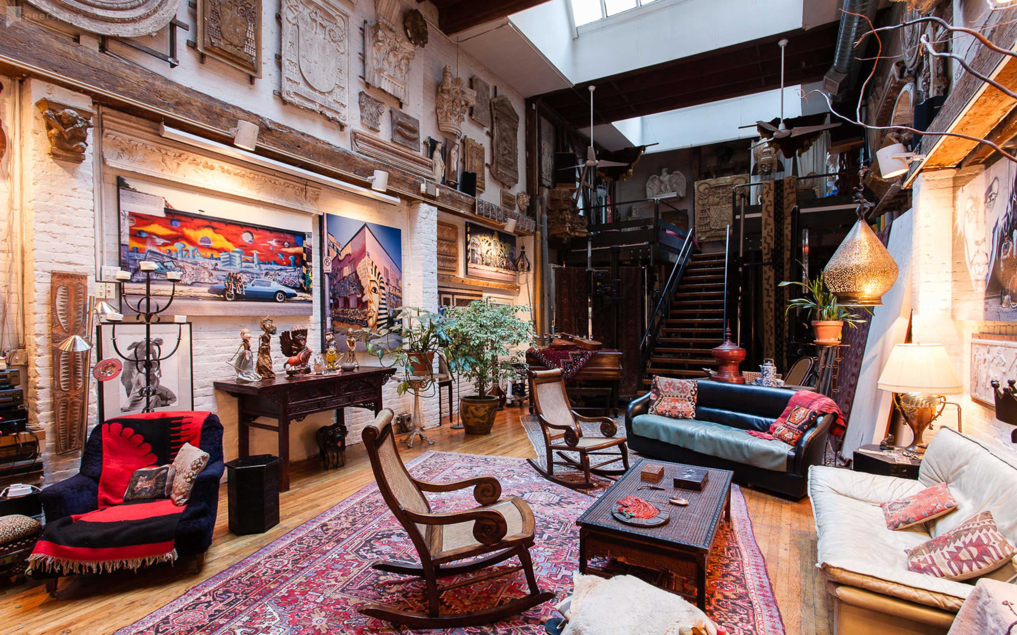 A spacious living room featuring eclectic artwork and decorative items, with a mix of seating options and a staircase in the background.