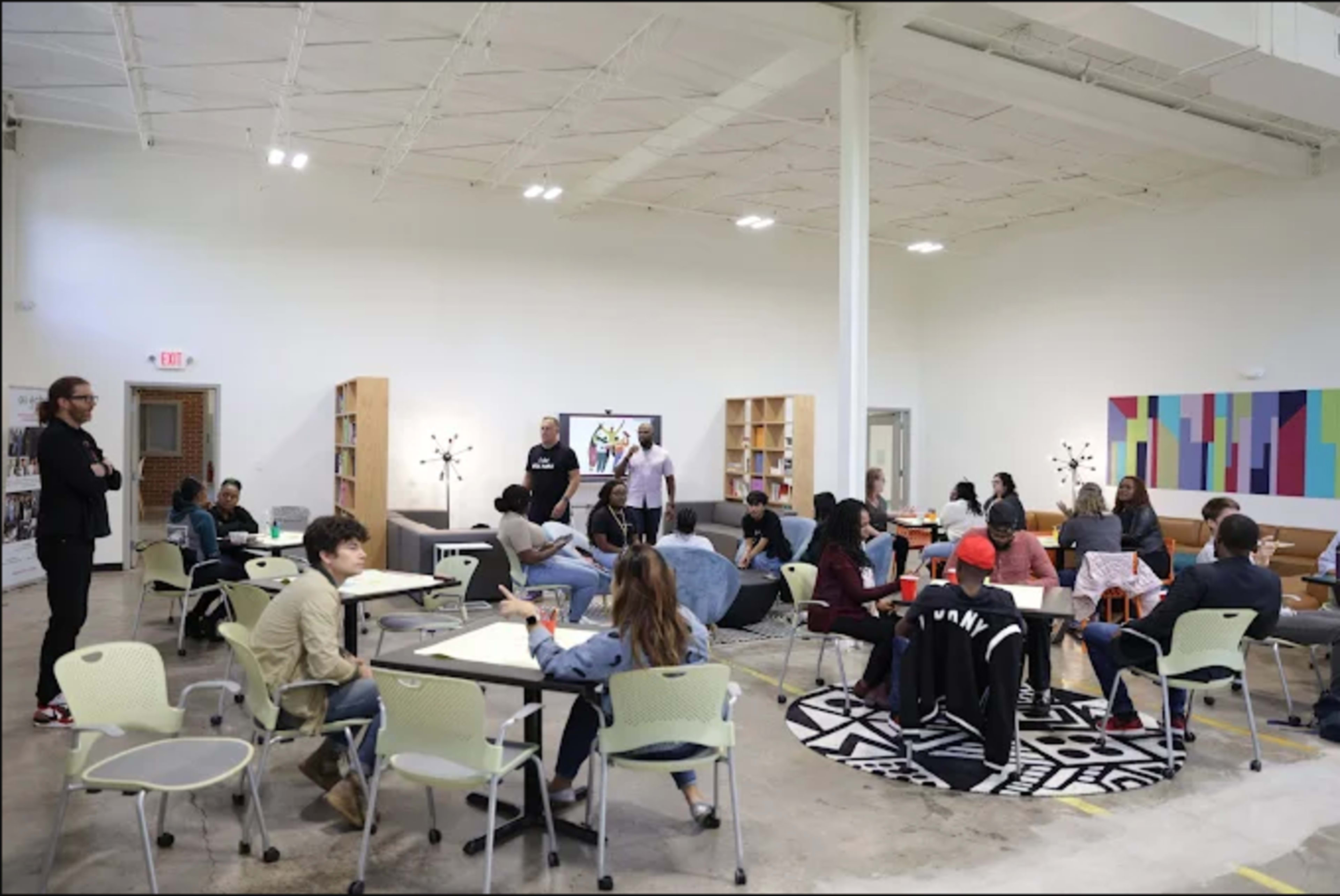 A group of people is seated in a large, bright room equipped with tables and chairs, while a few individuals stand at the front, engaging with the audience.