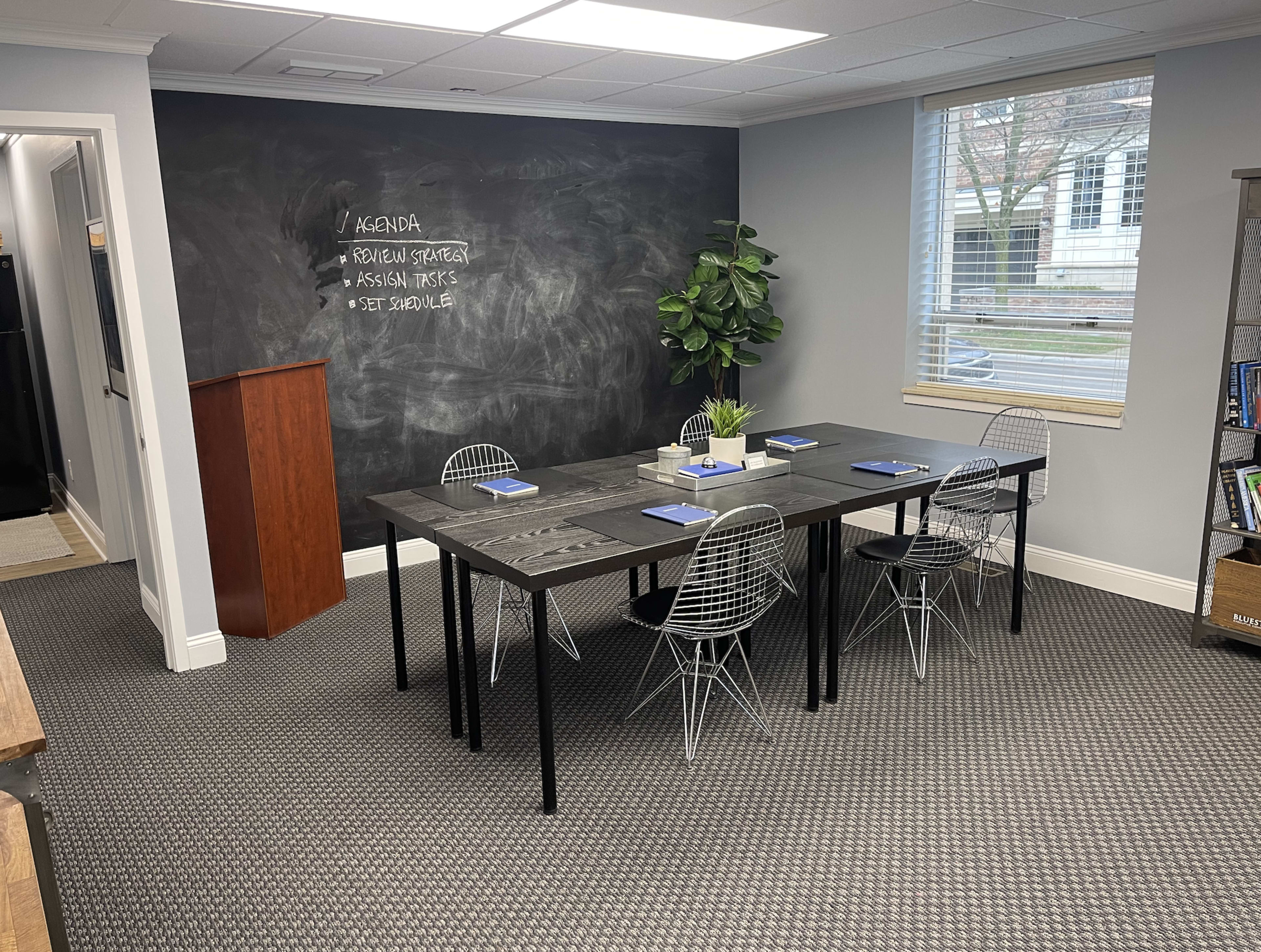 A modern conference room features a large black table surrounded by wire chairs, with a chalkboard wall listing an agenda and a potted plant in the corner.