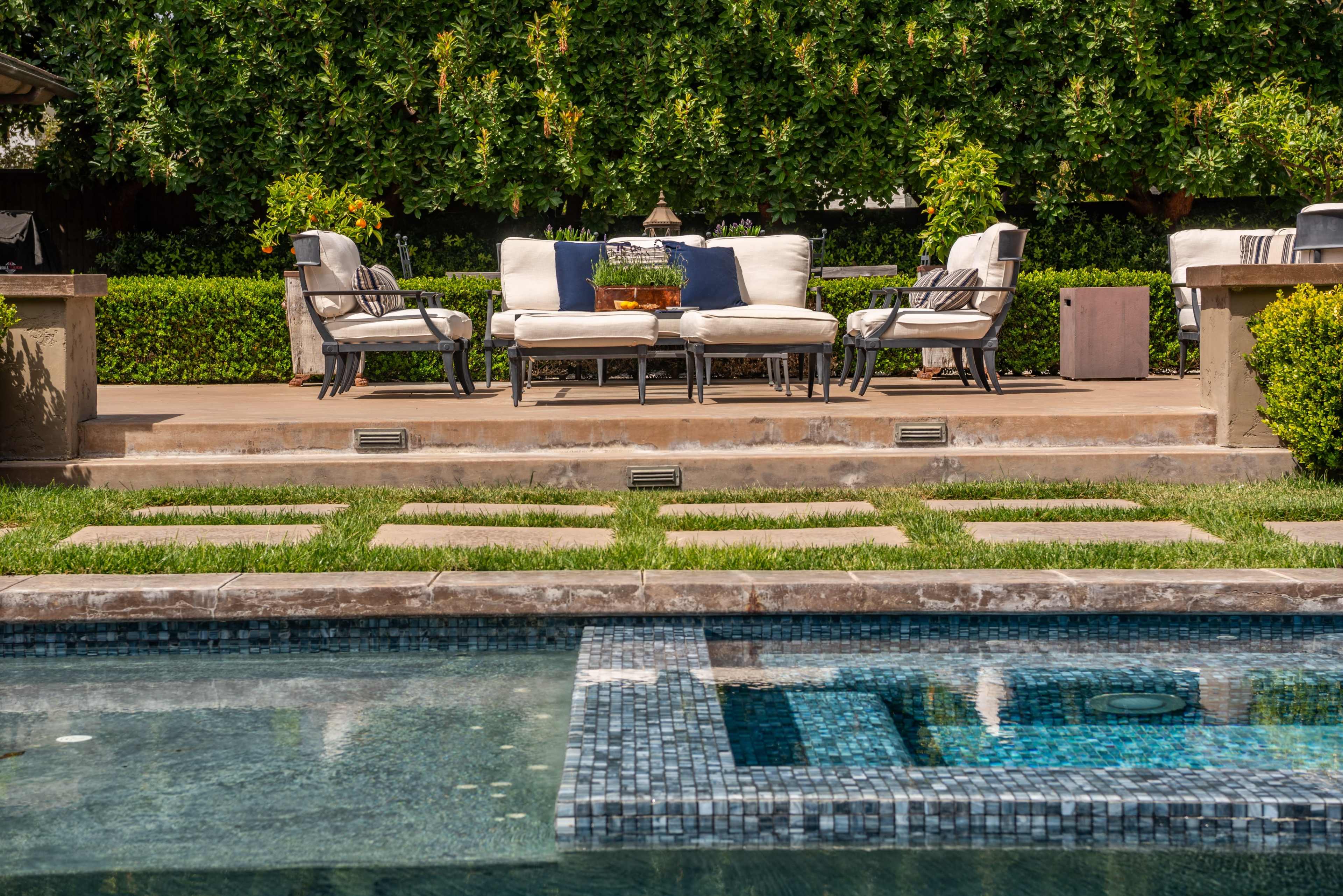 A patio area with outdoor furniture is positioned next to a swimming pool and surrounded by well-maintained greenery.