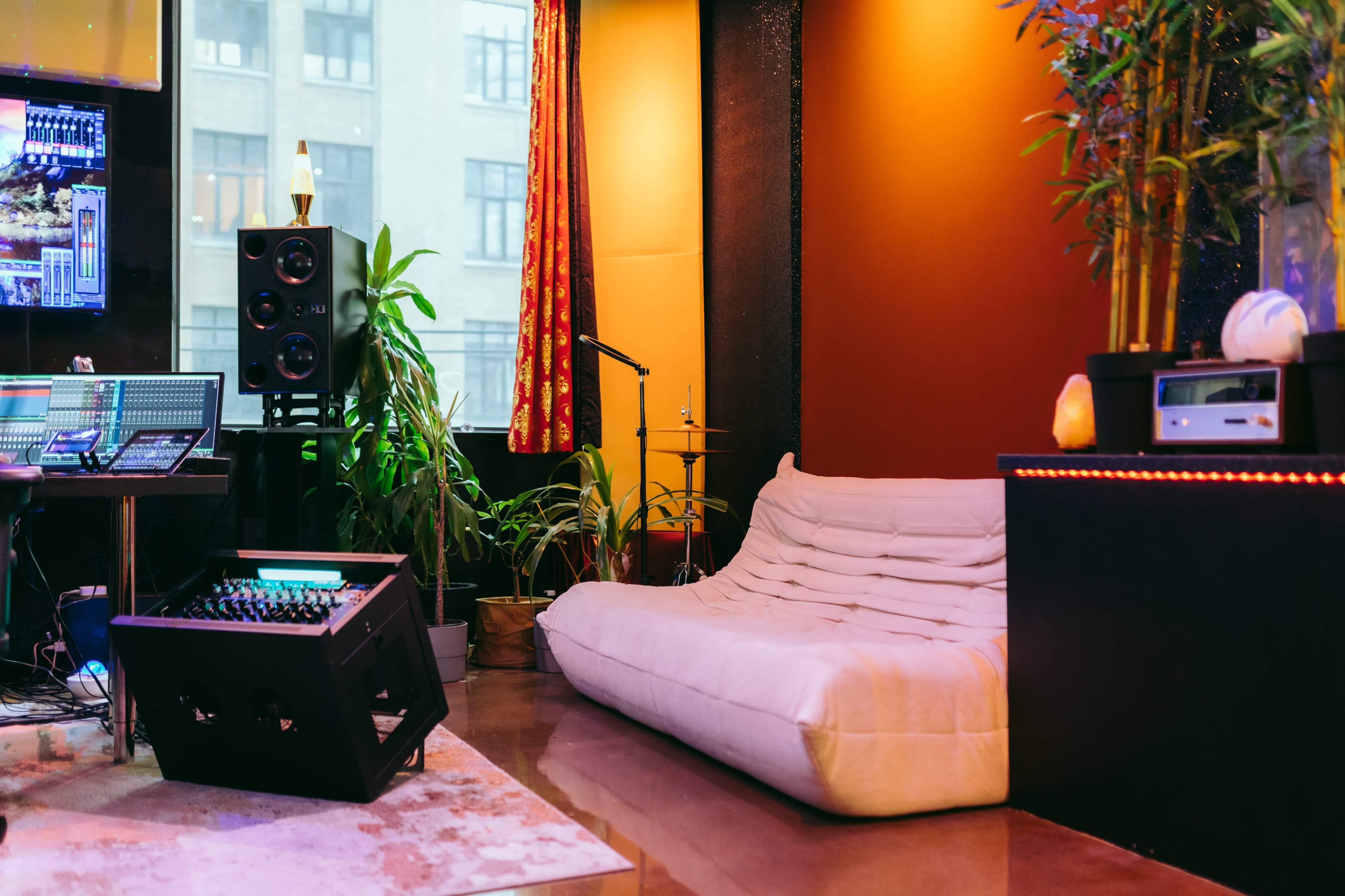 The image shows a cozy music studio with a white sofa, plants, a microphone stand, and audio equipment against a backdrop of large windows.