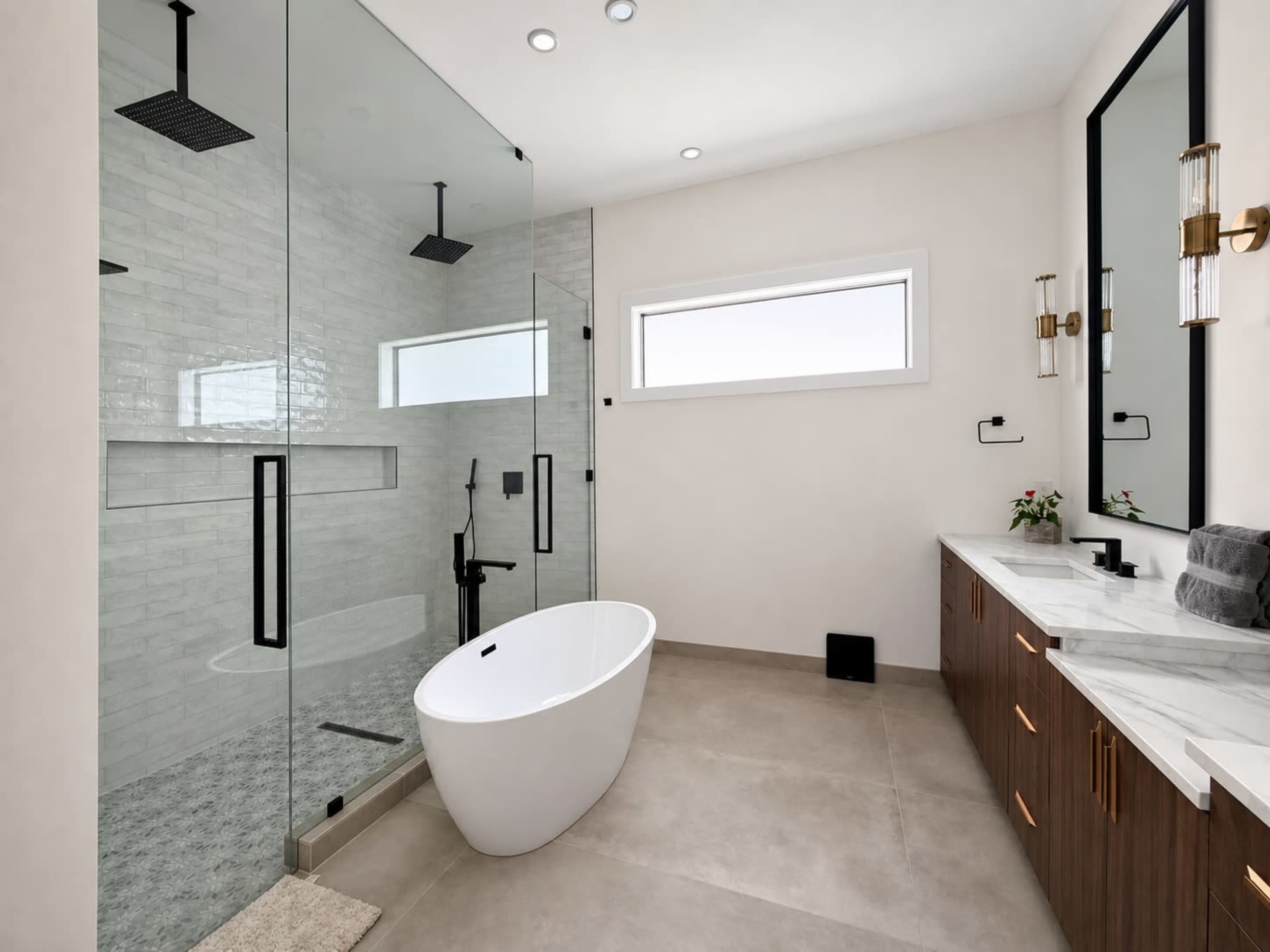The image shows a modern bathroom featuring a glass-enclosed shower, a freestanding bathtub, a double vanity with a marble countertop, and natural light coming through a large window.