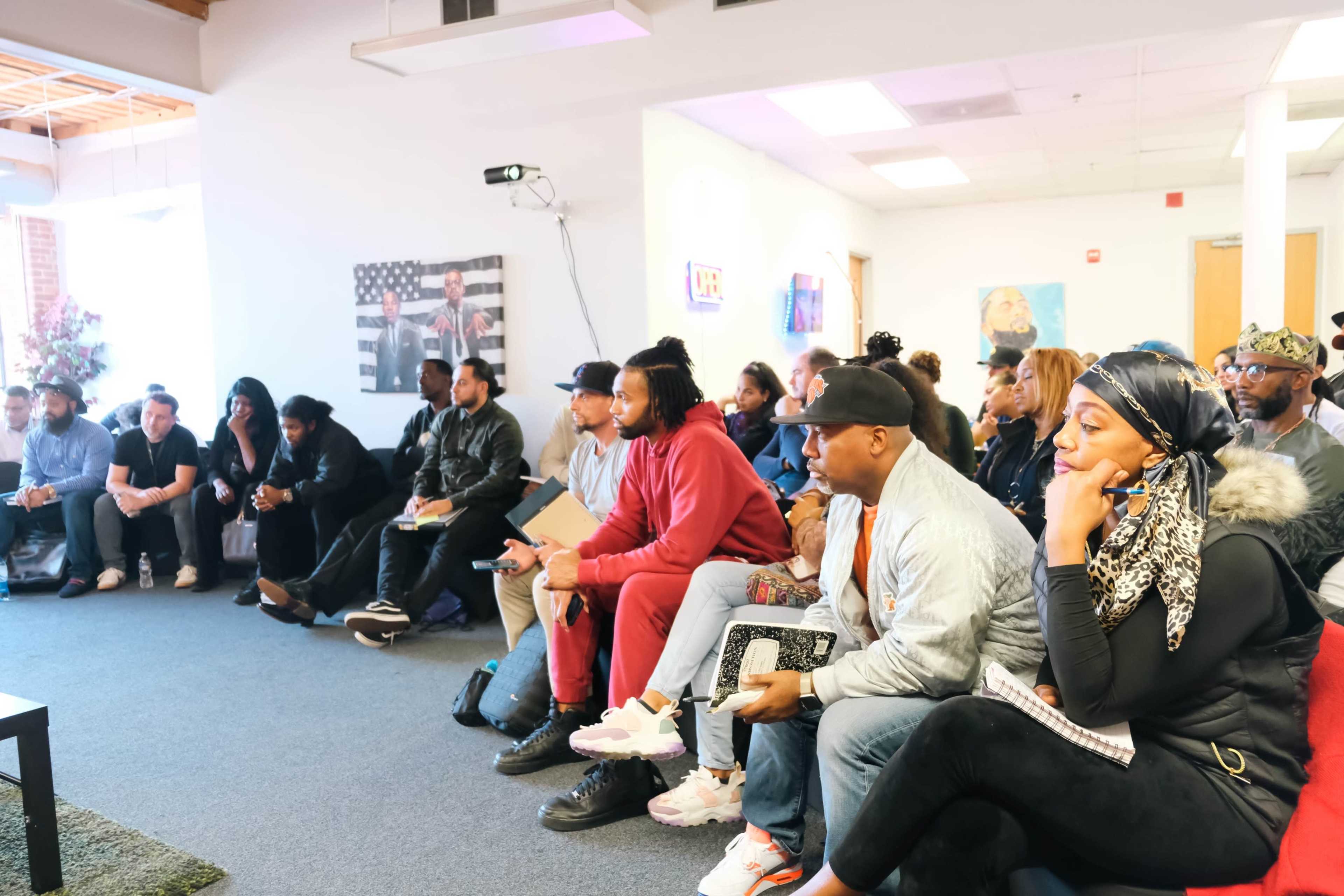 A diverse group of people sits in a well-lit room, focused on a presentation, with some holding notebooks and others watching a screen.