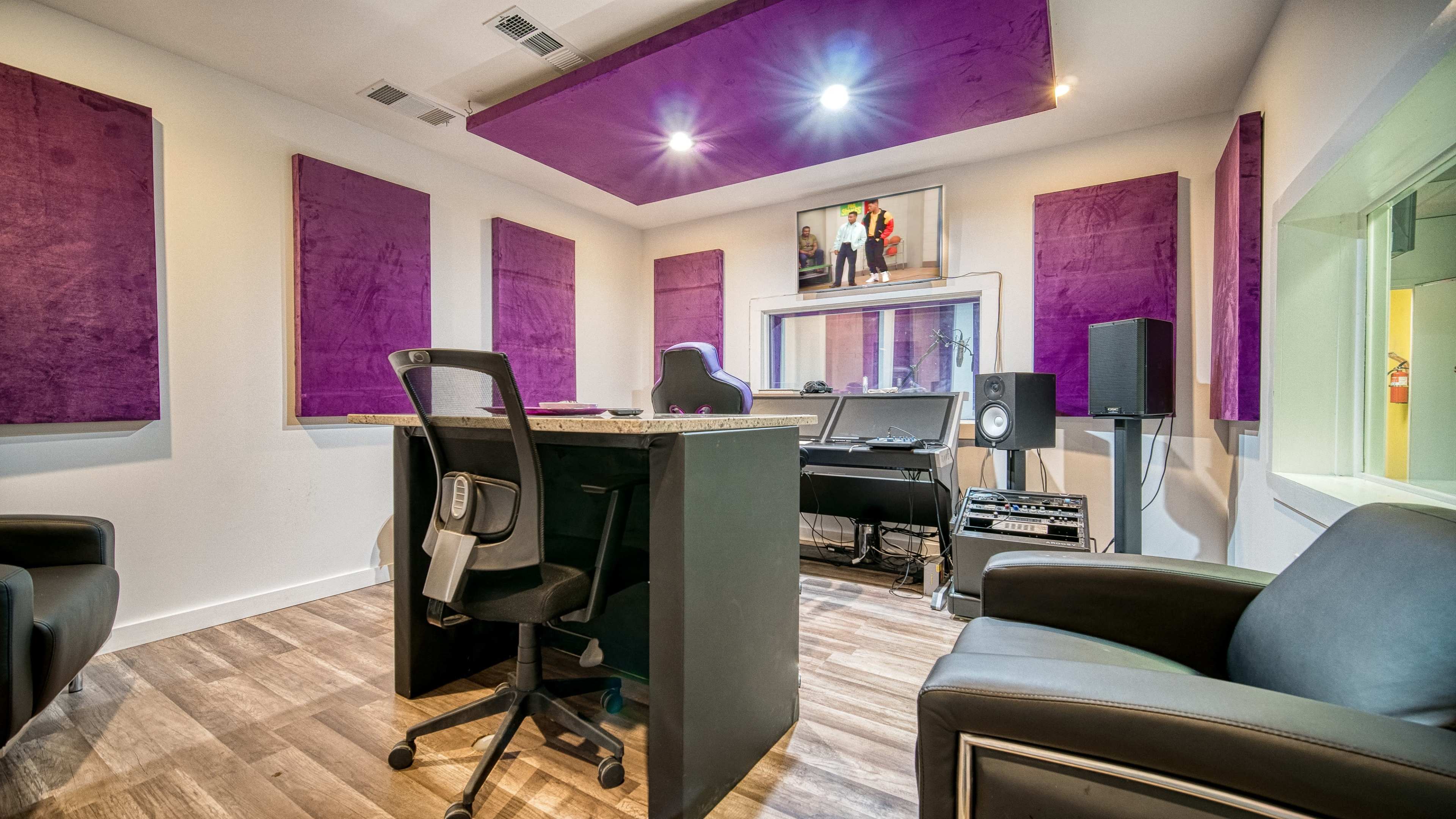 A modern recording studio featuring soundproofed purple panels, a central desk with a computer setup, and leather chairs.