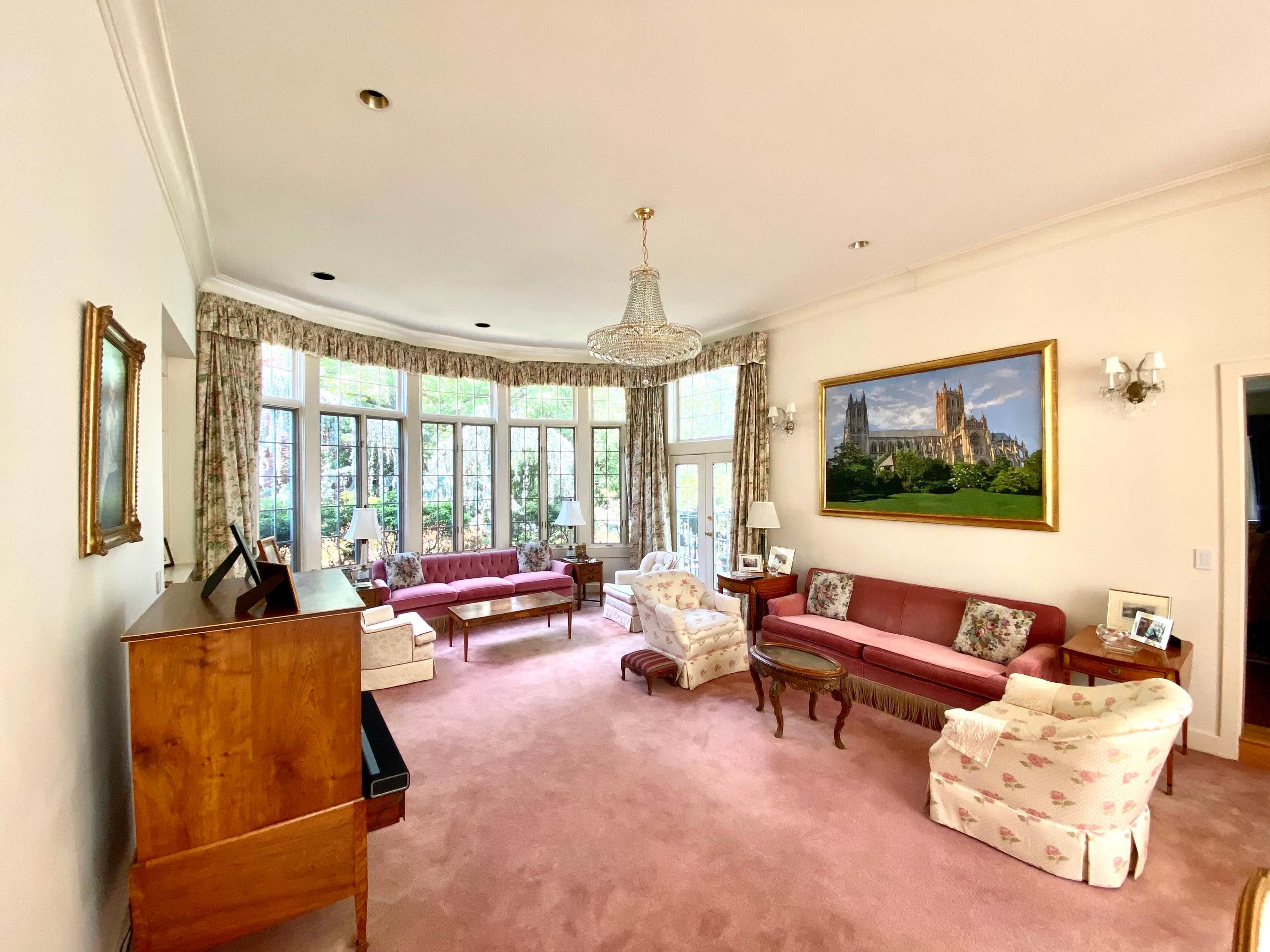 A spacious living room featuring two sofas, a patterned armchair, a wooden piano, and large windows with views of a garden.
