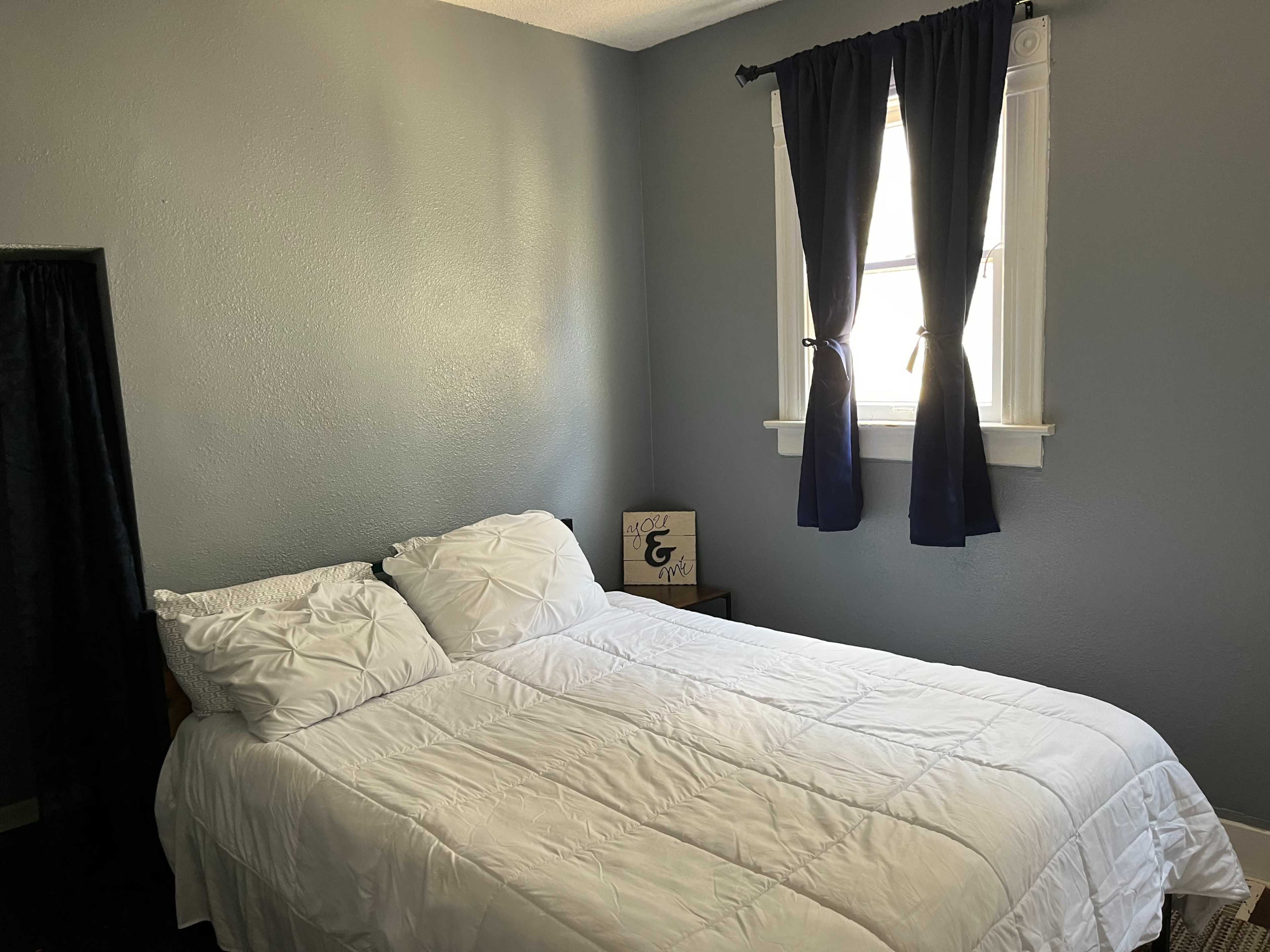 A simple bedroom with a bed covered in a white comforter and two pillows, a window with dark curtains, and a small decorative item on a nightstand.