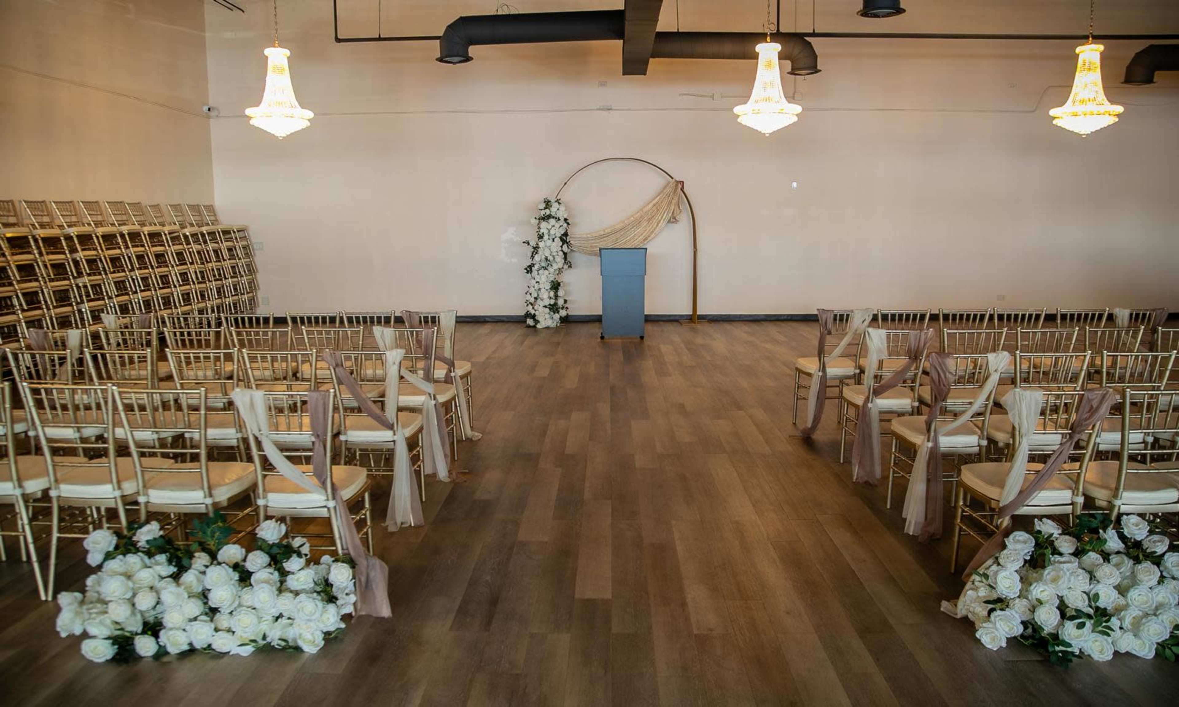 The image shows a spacious event hall with rows of gold chairs, a podium at the front, and floral decorations on the floor.