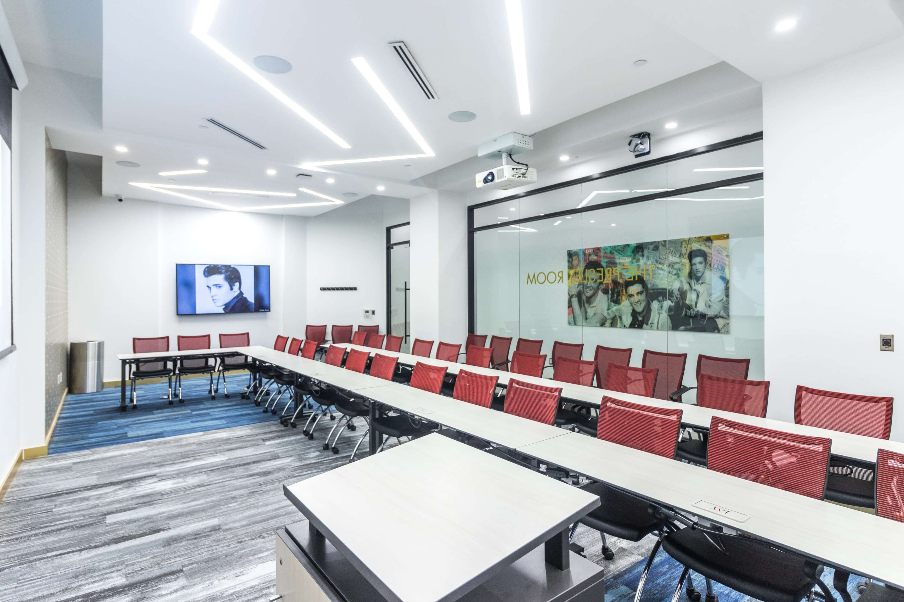 The image shows a modern conference room with a long table, red chairs, a projector, and a large screen displaying a black-and-white photo on one wall.