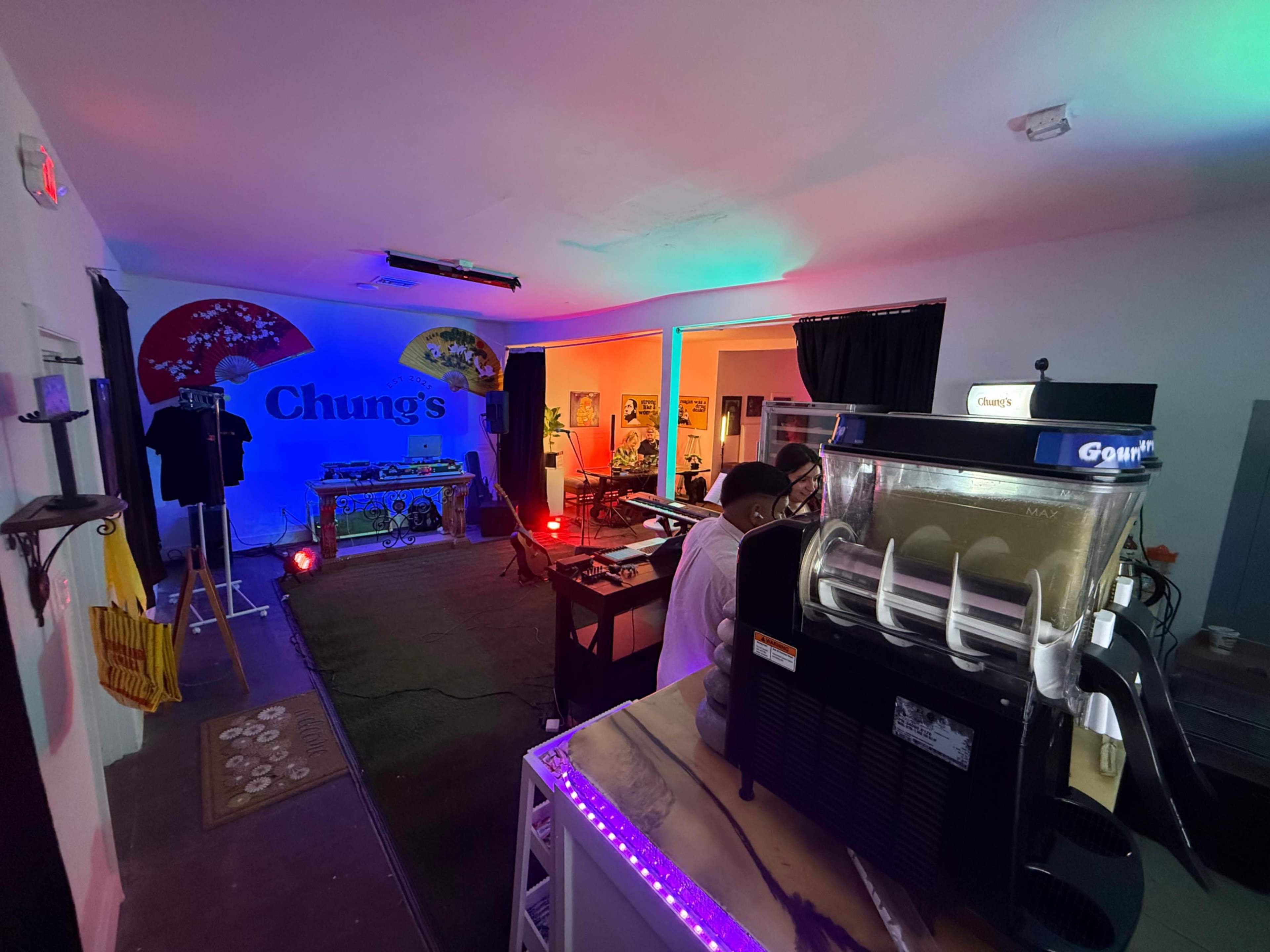 The image shows a brightly lit room with a slushy machine in the foreground, and a stage setup in the background featuring colorful lighting and decorations.