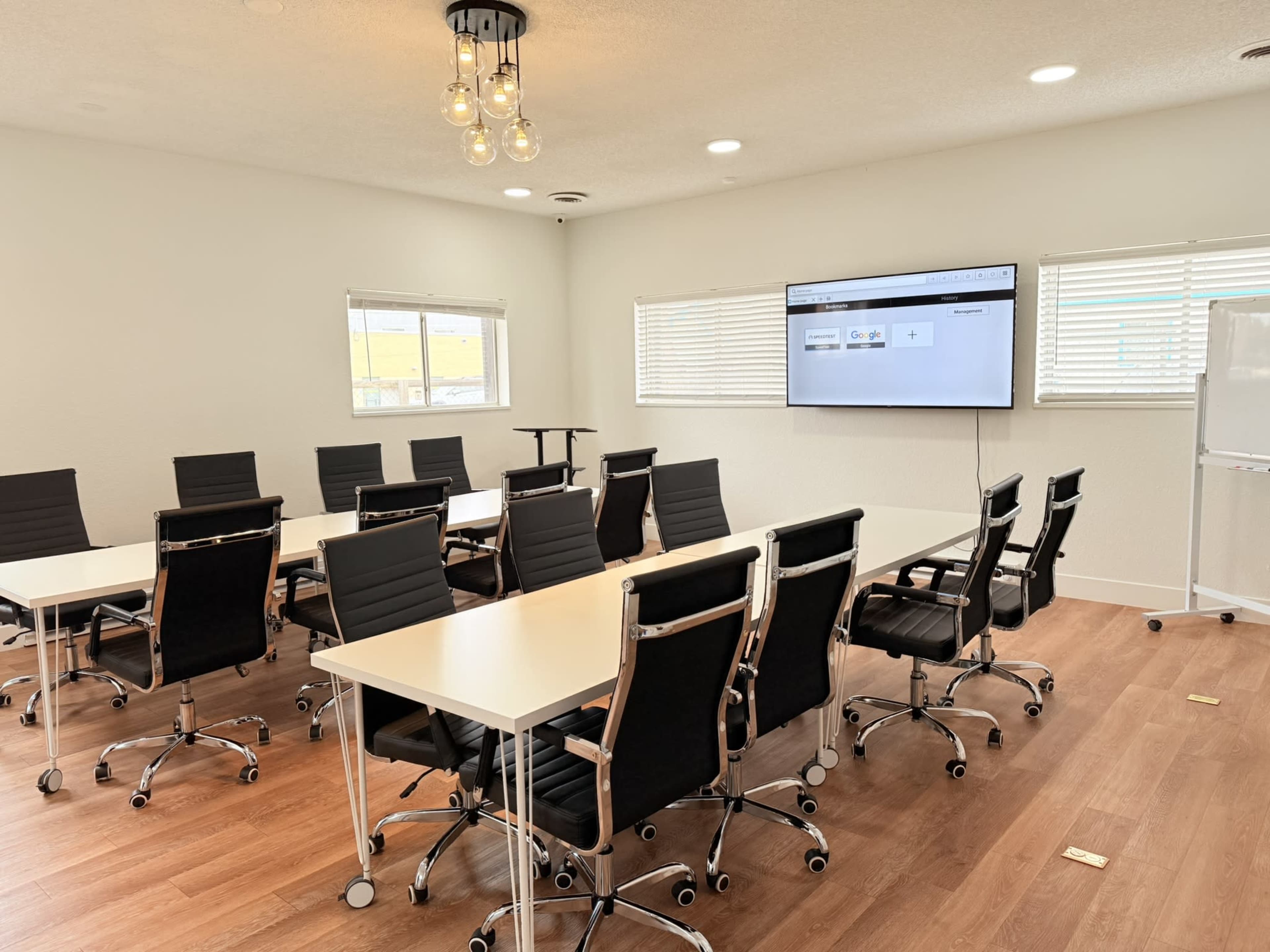 A modern conference room features several black office chairs arranged around white tables, with a large screen mounted on the wall.
