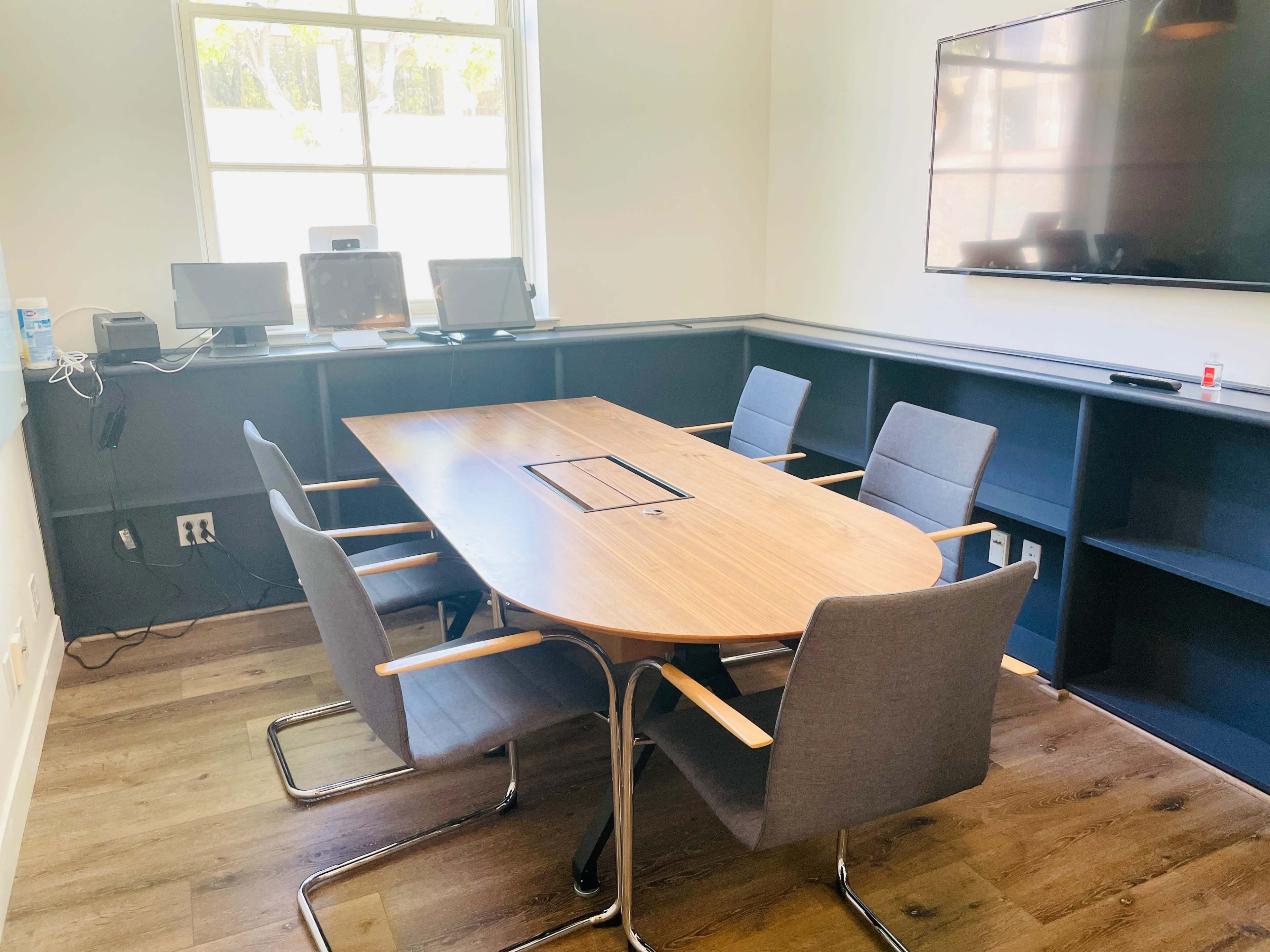 A modern conference room featuring a wooden table surrounded by five gray chairs, with a wall-mounted television and two computers on a desk in the background.