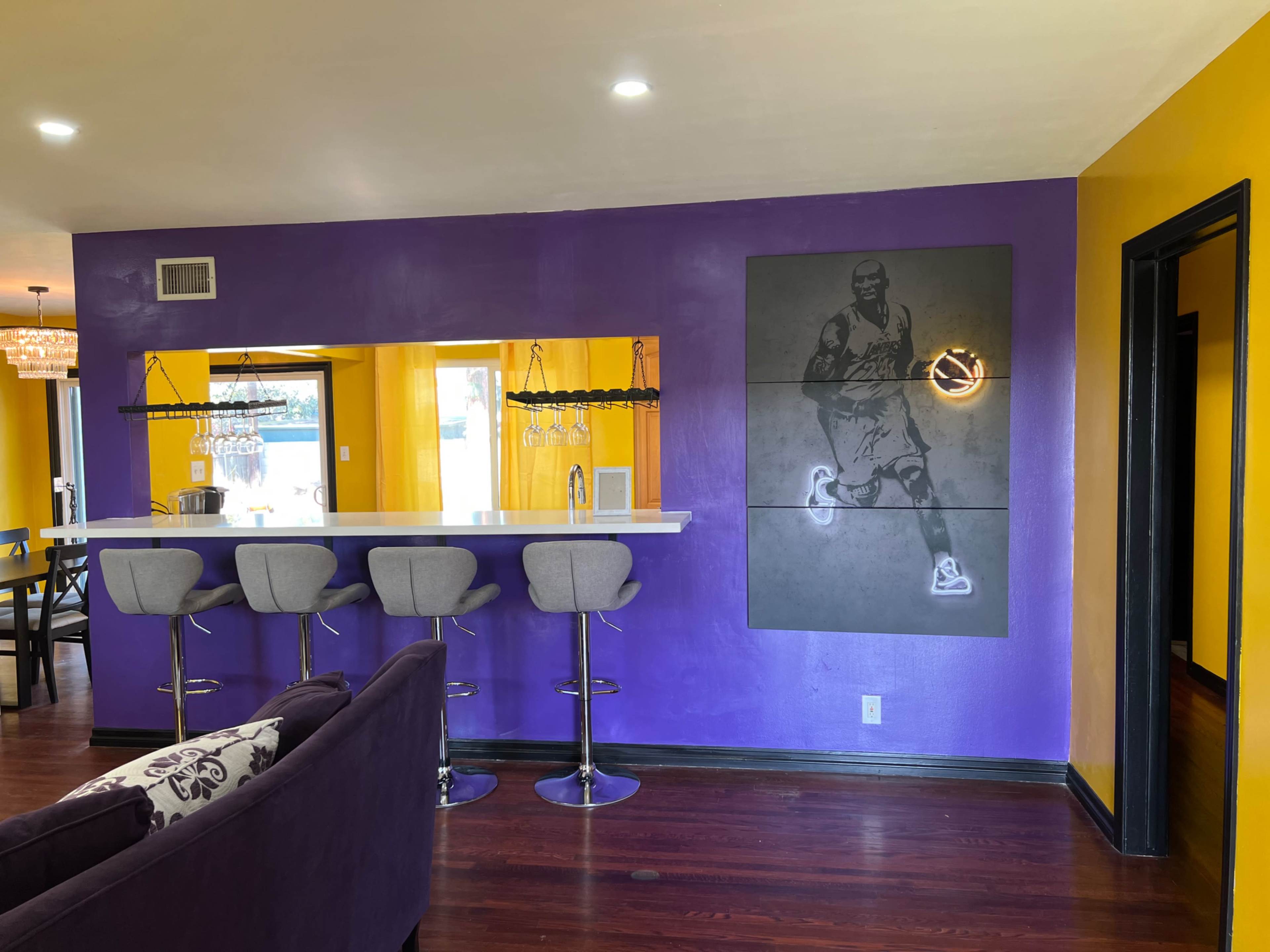 A brightly colored room with purple and yellow walls, a bar with gray stools, and a basketball-themed artwork on the wall.