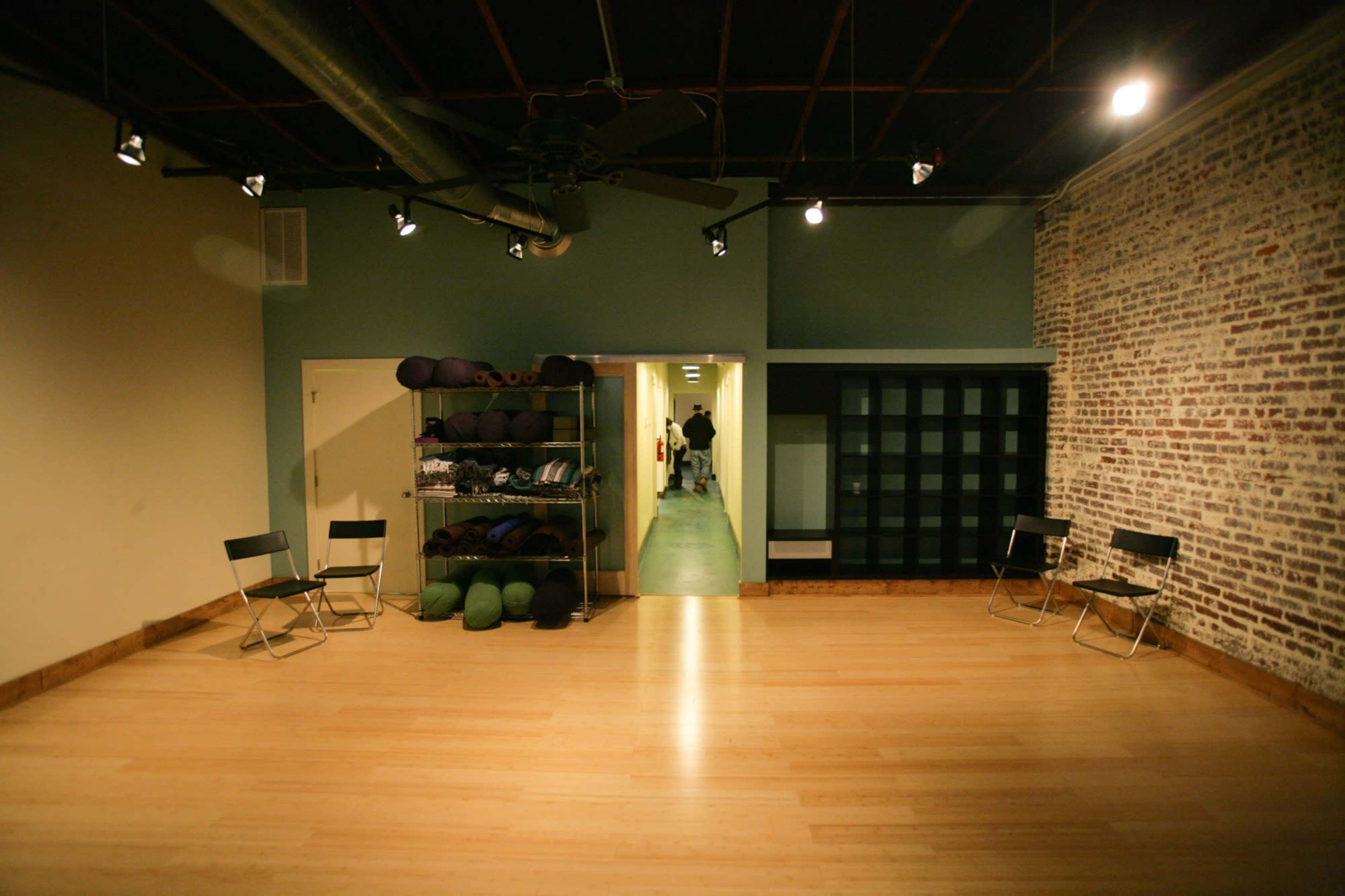 A spacious, empty room with wooden floors, a stack of yoga mats on the side, and a hallway leading to another area.