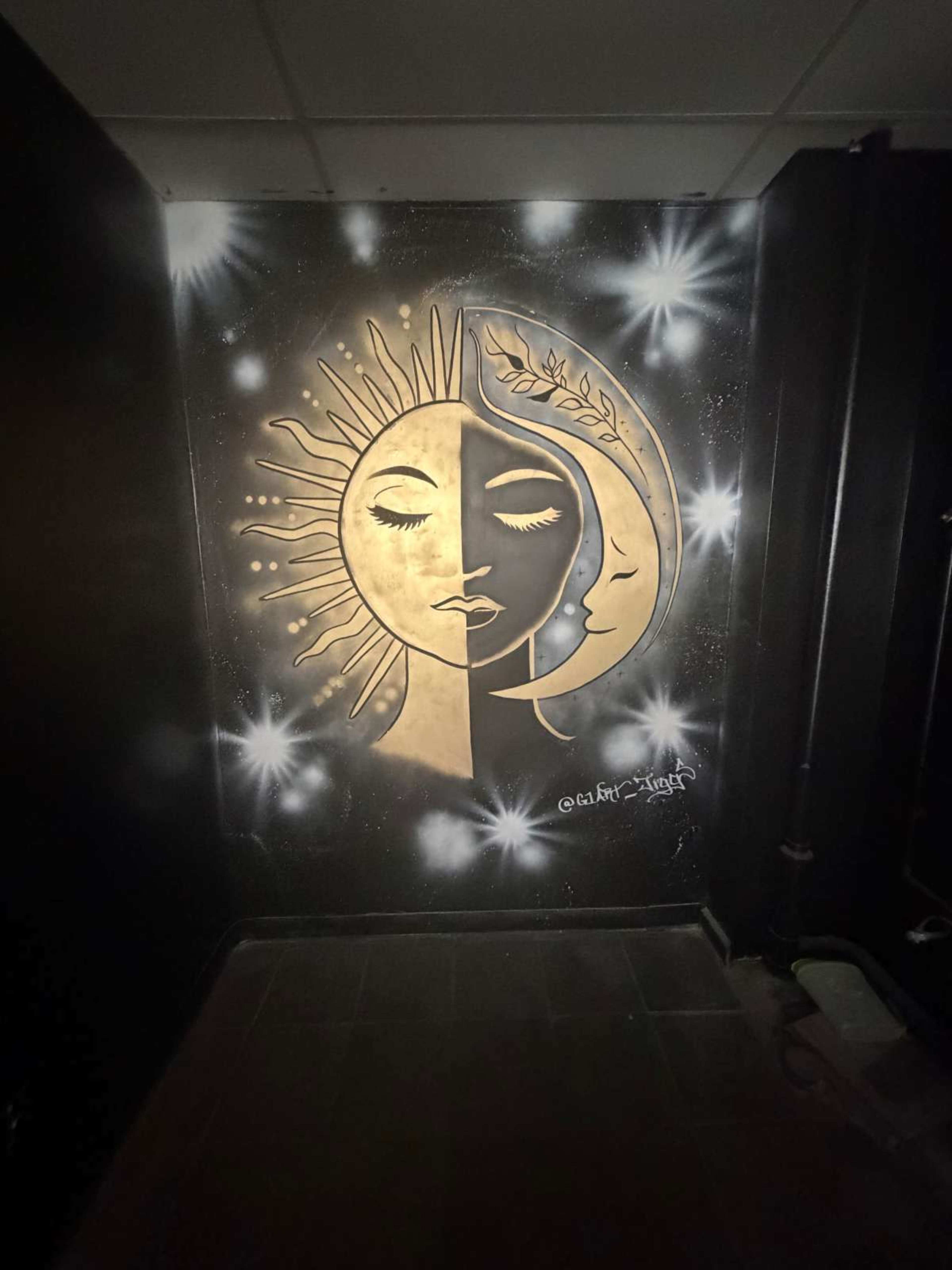 The image shows a mural on a black wall featuring a design of a sun and moon with intricate details, surrounded by stars.