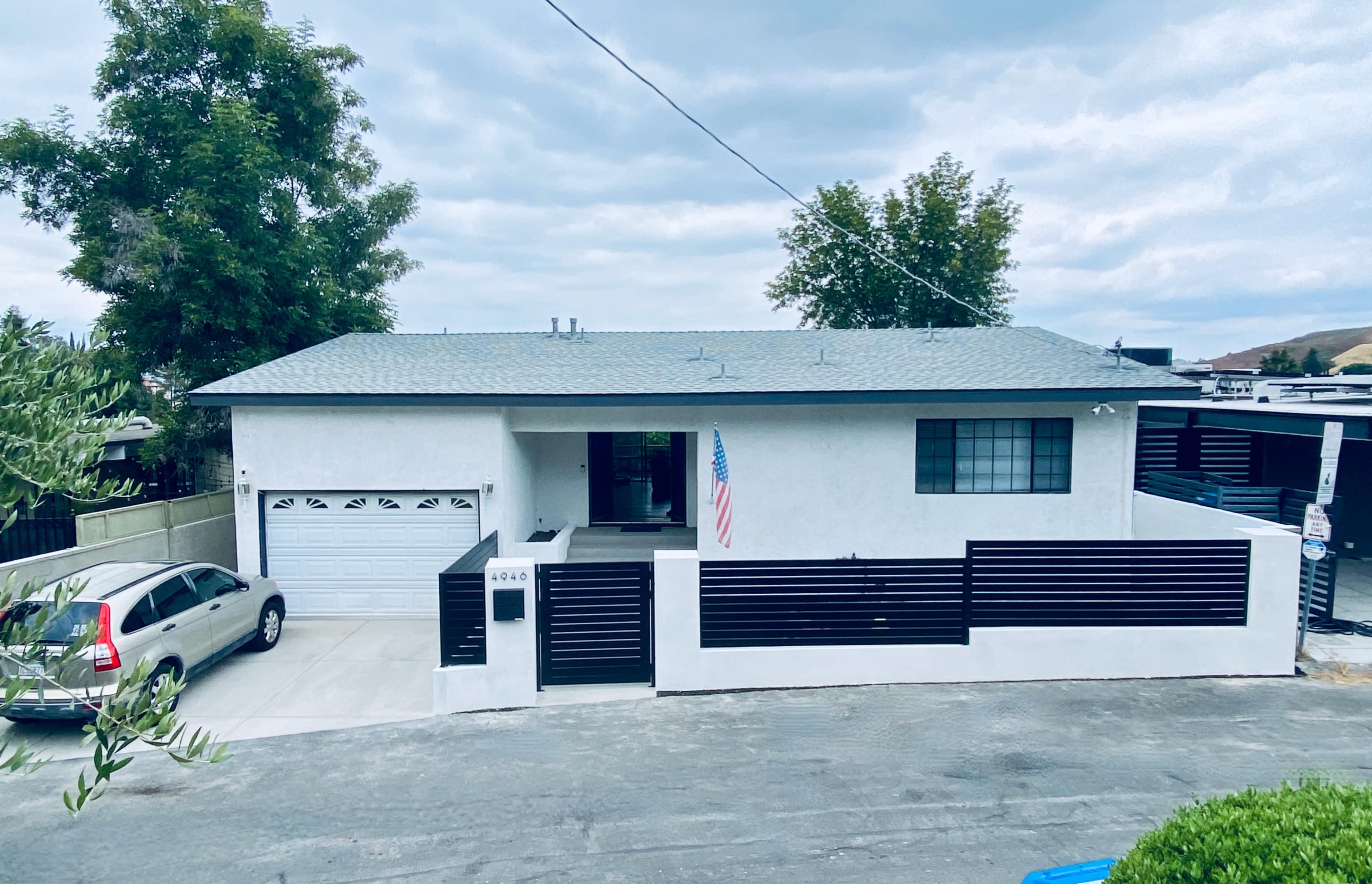 The image shows a single-story white house with a gray roof, a garage, and a front yard fenced with horizontal black slats, along with an American flag displayed near the entrance.