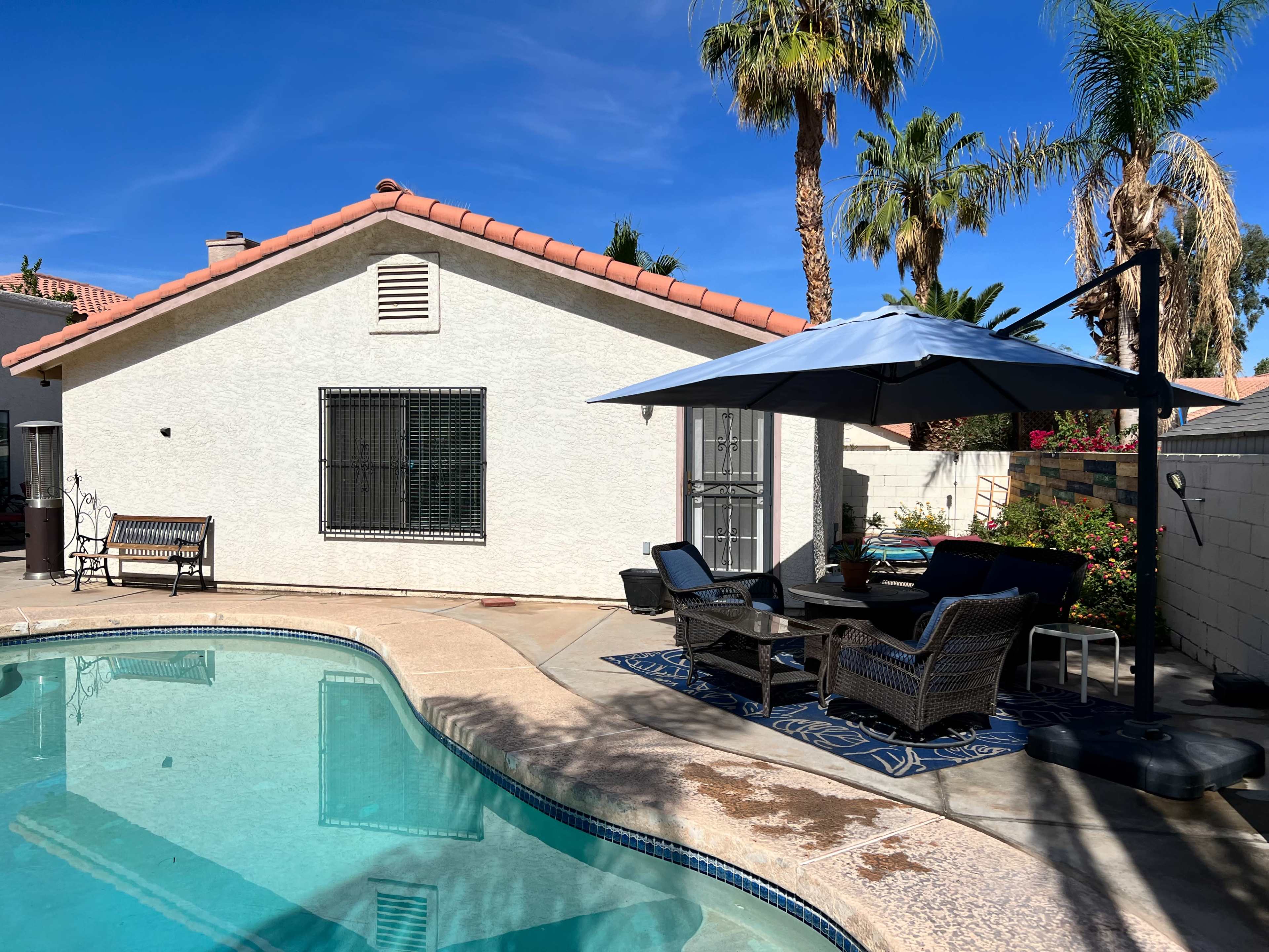 A backyard scene features a swimming pool bordered by a patio area with seating and an umbrella, surrounded by palm trees and a white house.
