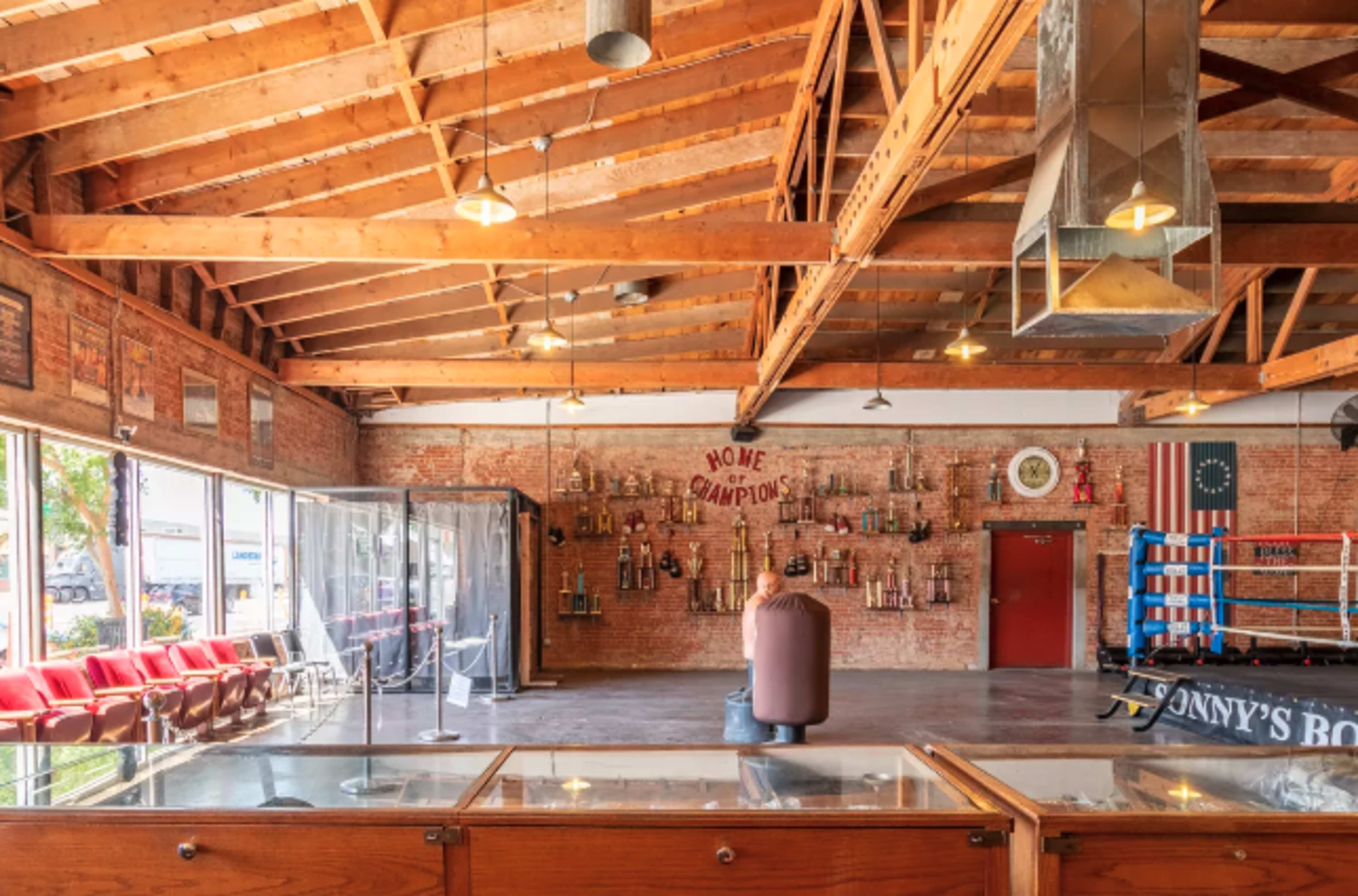 The interior of a boxing gym features exposed wooden beams, brick walls adorned with trophies, and a boxing ring in the corner.