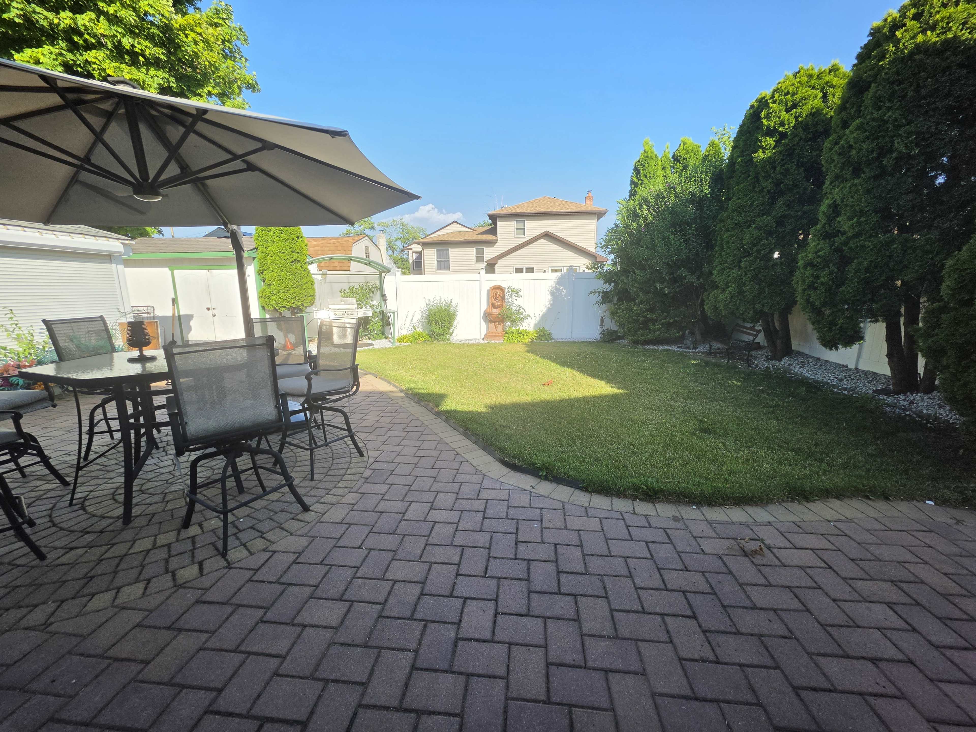 The image shows a backyard patio with a dining set under an umbrella, a grassy area, and tall hedges against a white fence.