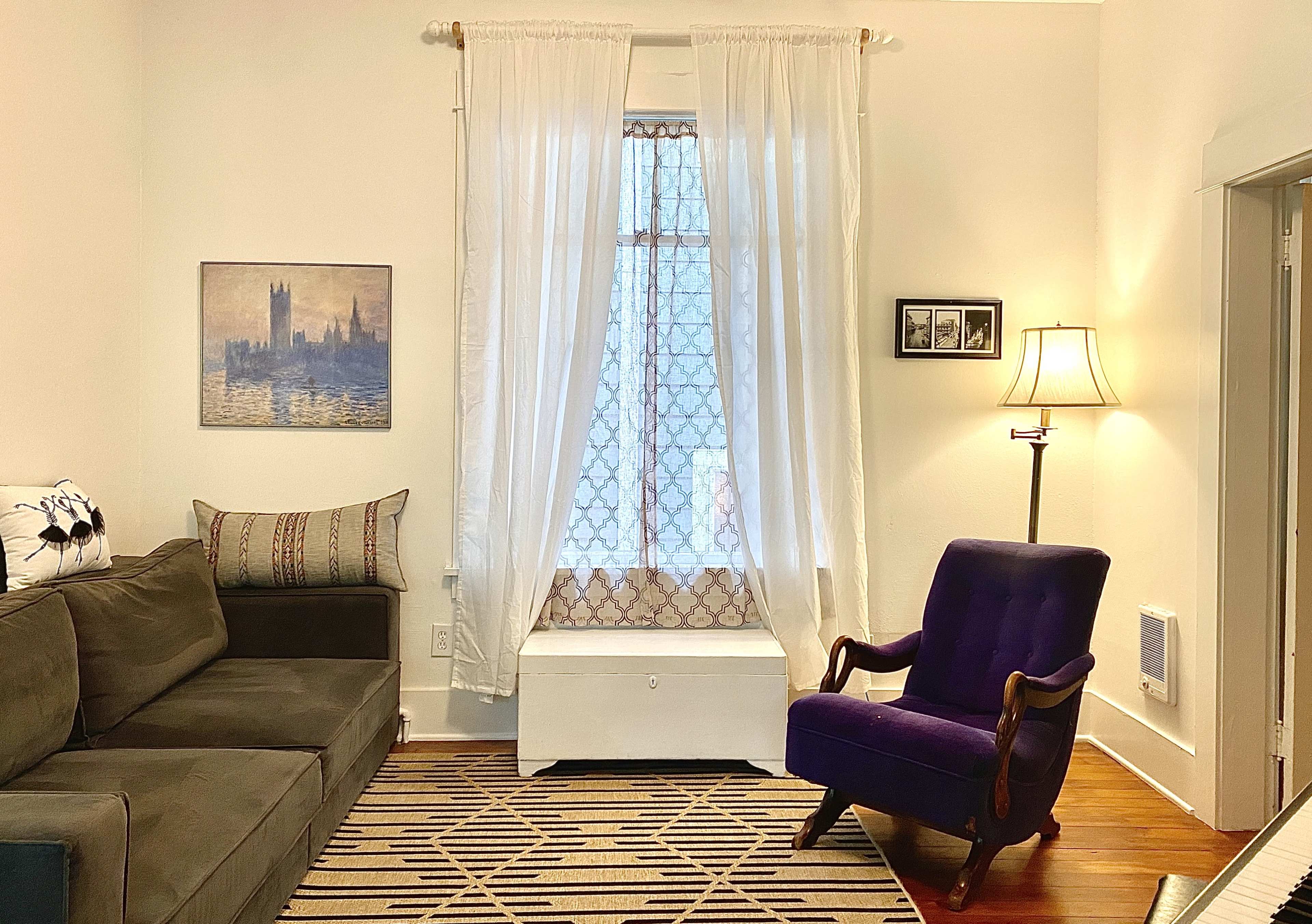 A cozy living room features a gray sofa, a purple armchair, and a window draped with white curtains, illuminated by a table lamp.