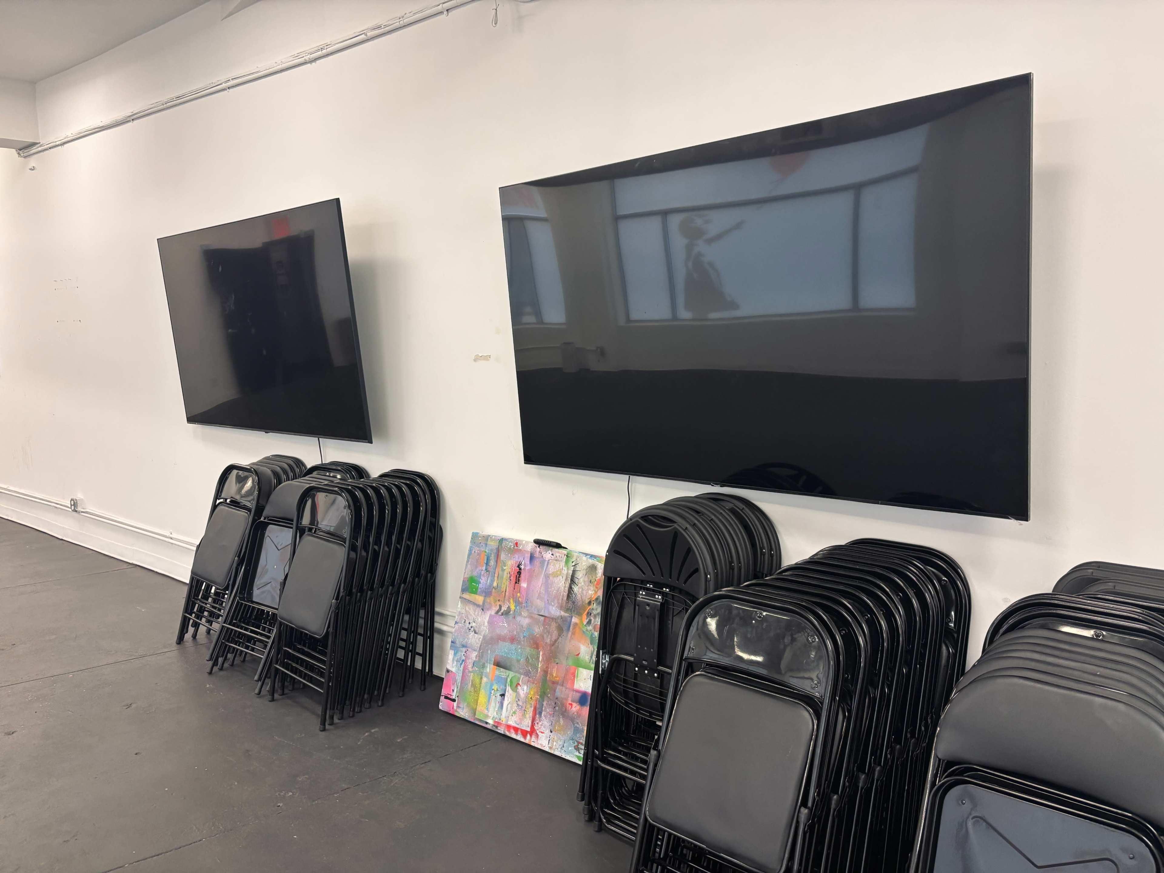 The image shows two large flat-screen televisions mounted on a white wall next to a stack of black folding chairs and a colorful abstract painting leaning against the wall.