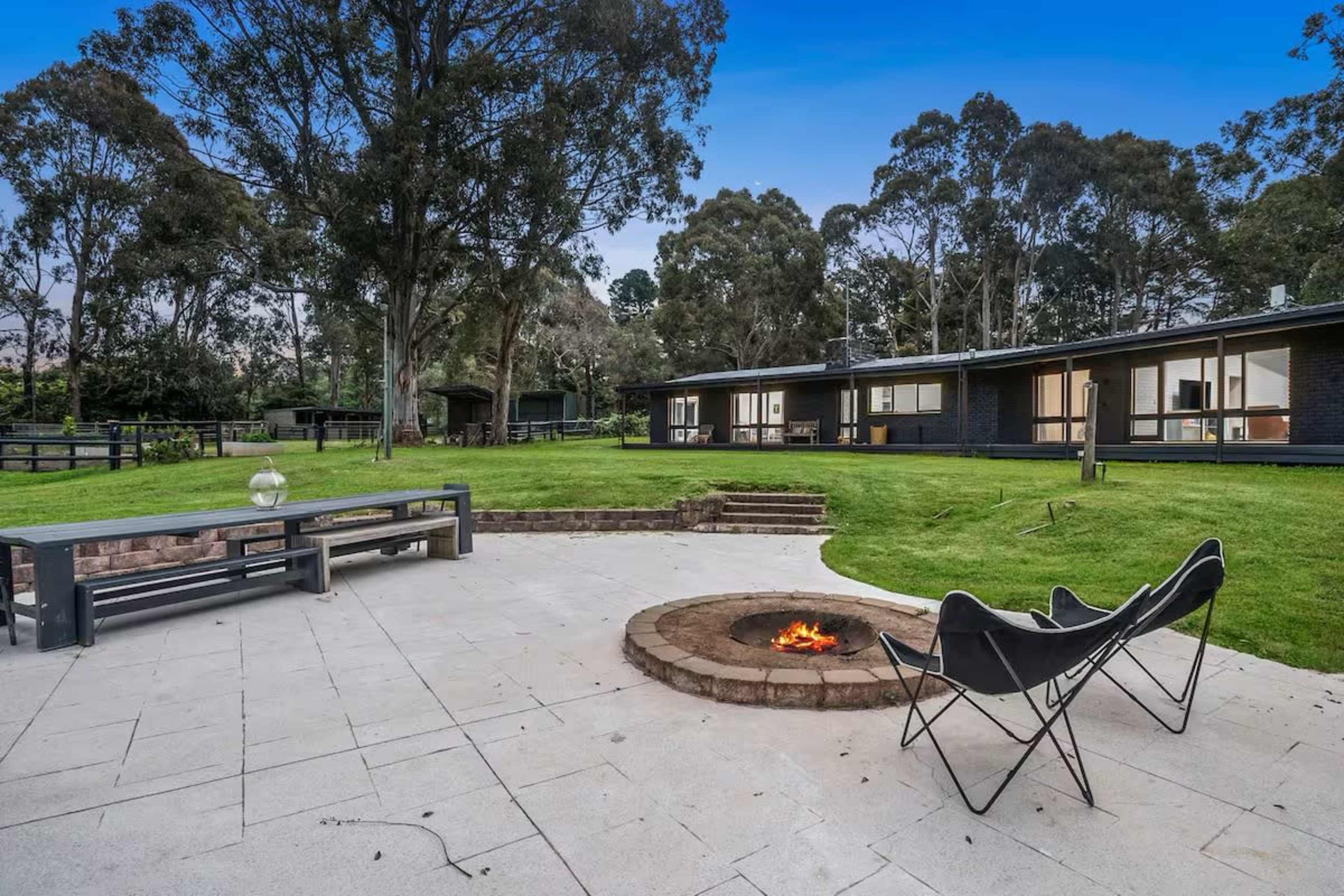 The image shows a backyard with a stone fire pit surrounded by chairs, a patio area, and a lush lawn with trees and a house in the background.