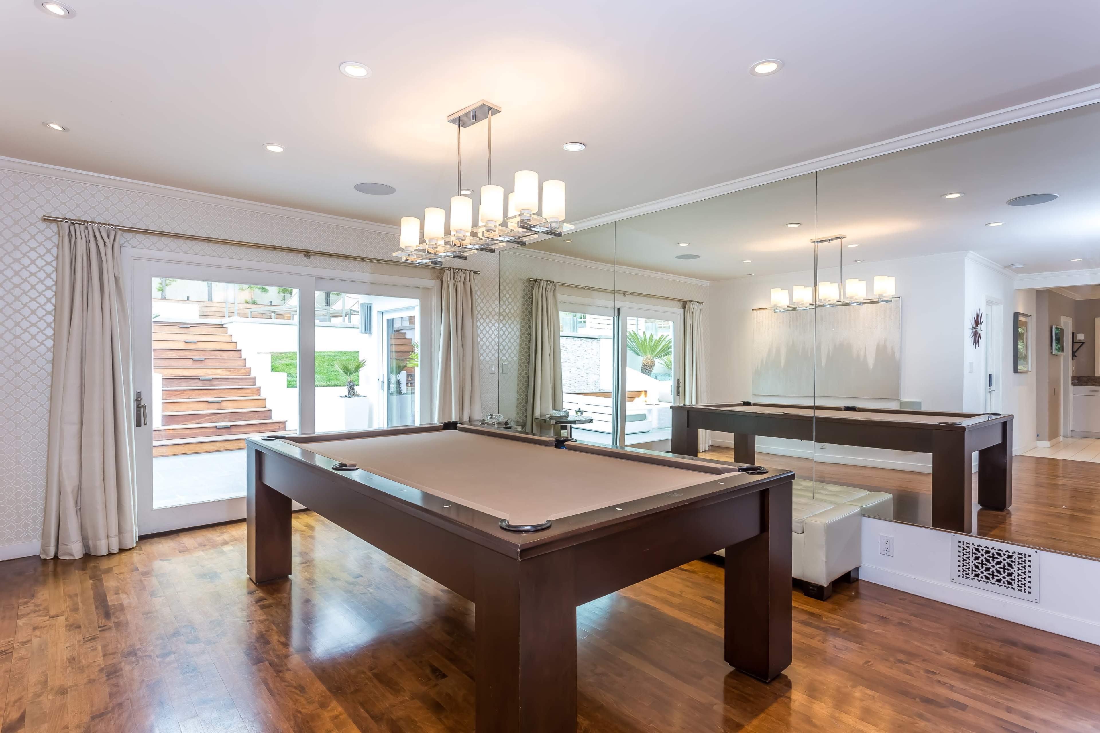 The image shows a well-lit game room featuring a billiard table, large mirrors, and sliding glass doors leading to an outdoor area.