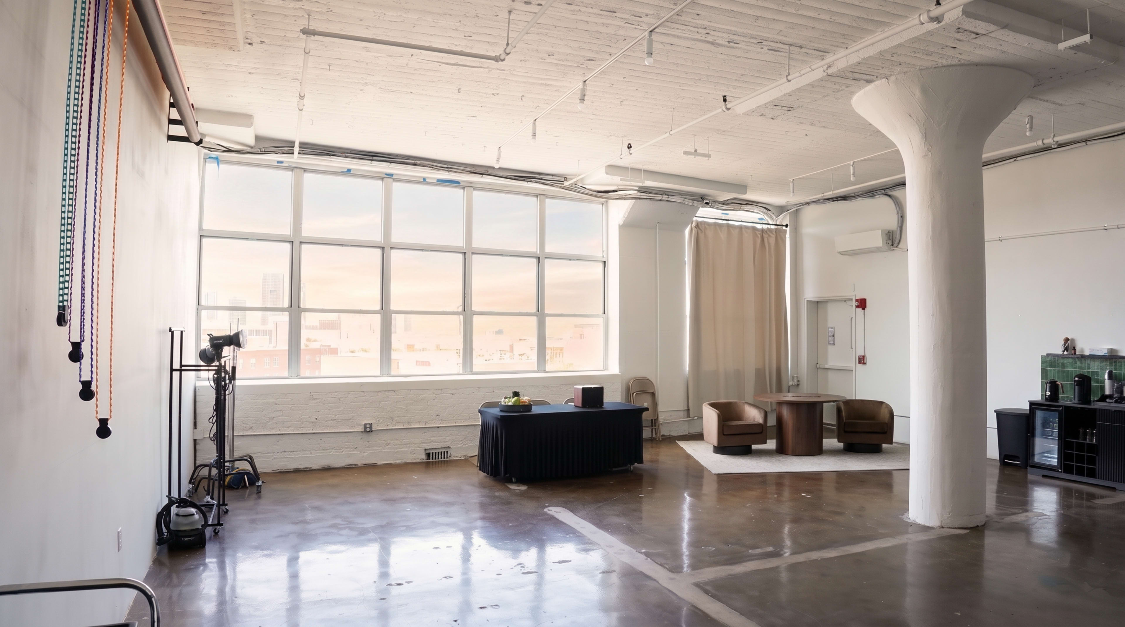 The image shows a spacious, industrial-style room with large windows, a seating area, and a few furnishings, including a table and some chairs.