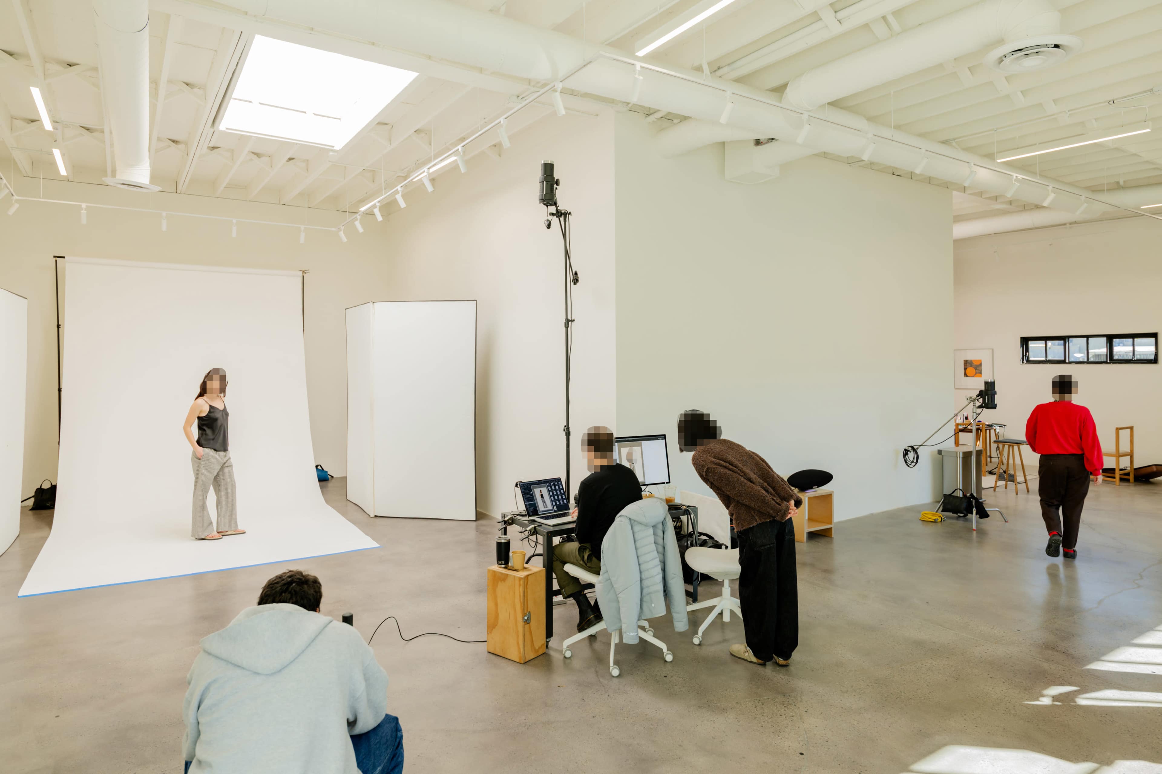 A model poses on a white backdrop in a spacious photo studio where several people are operating equipment at computers.