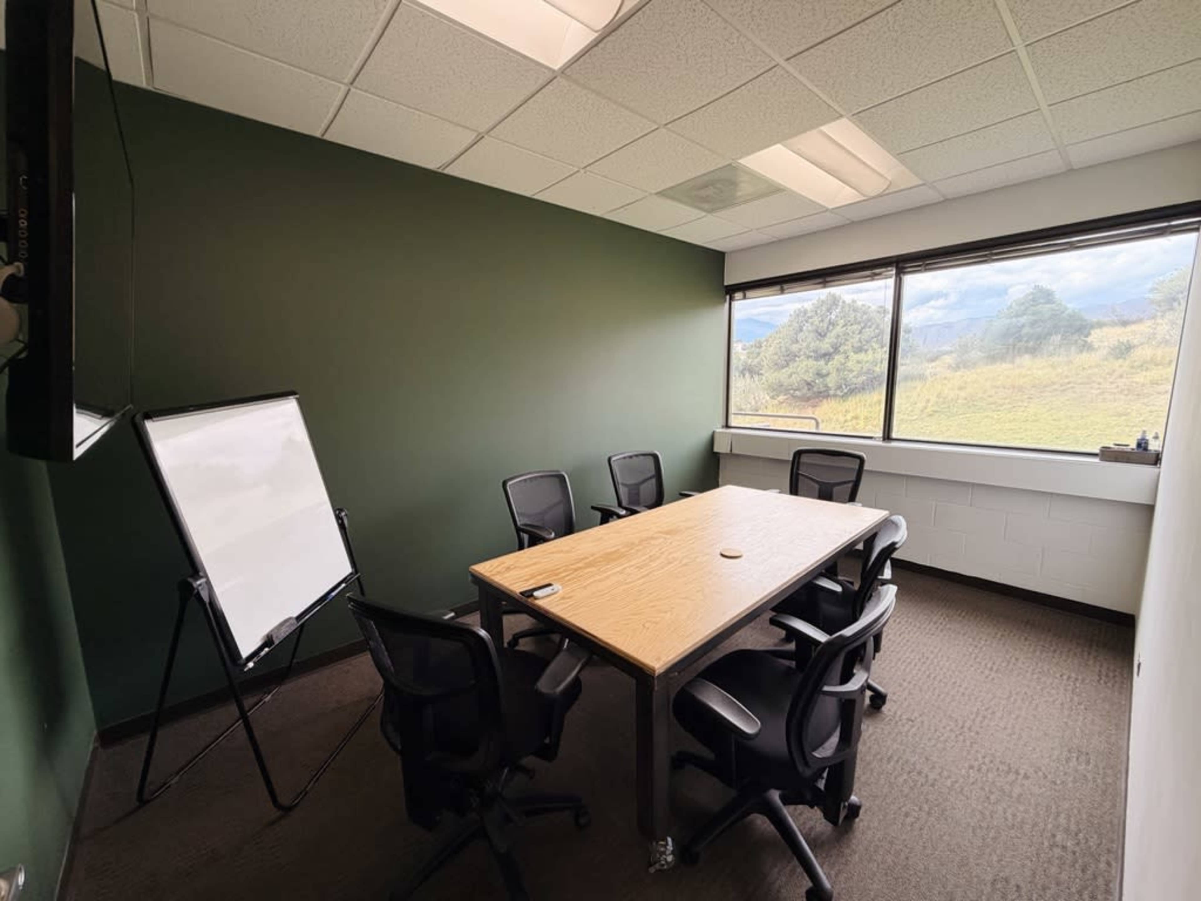 The image shows a small conference room with a wooden table surrounded by five black chairs, a whiteboard, and large windows offering a view of hills and trees.