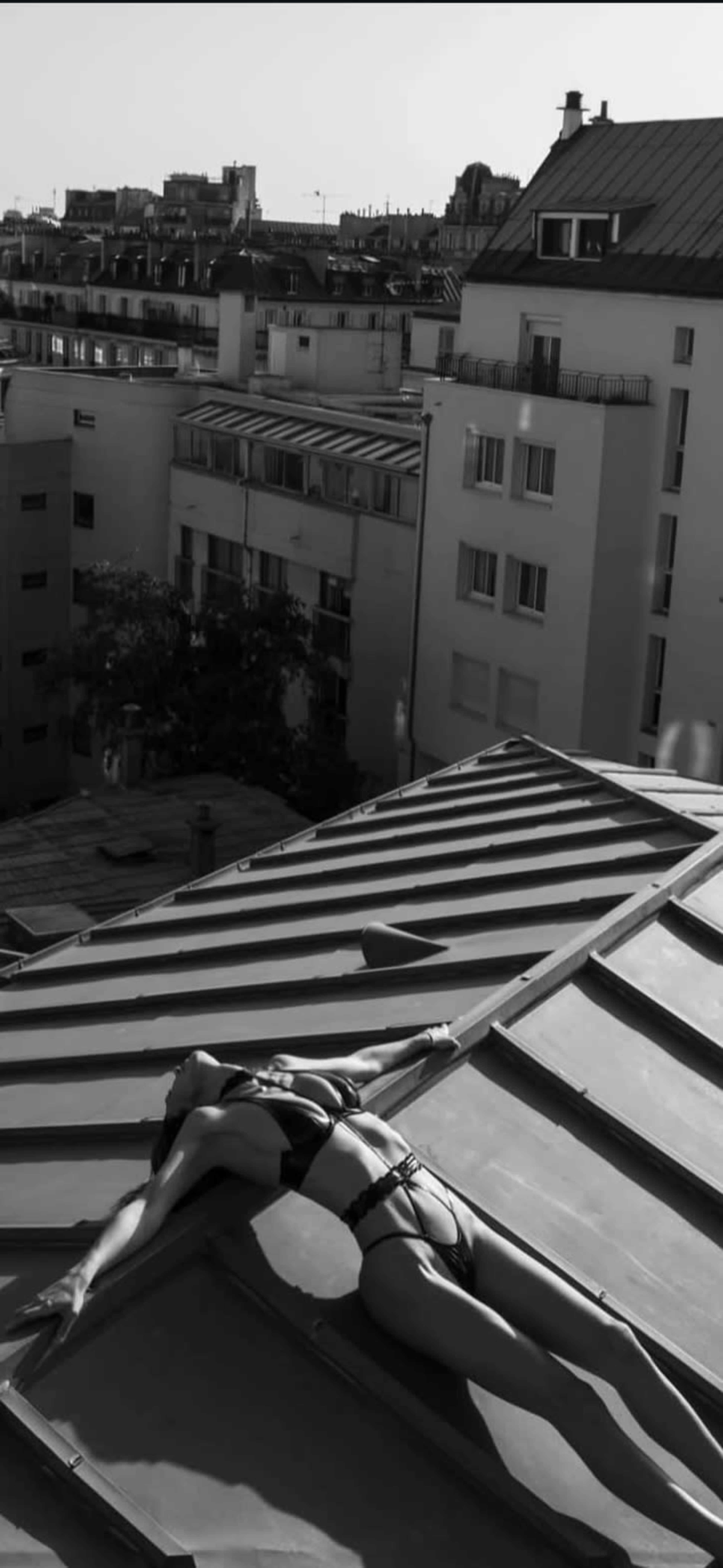 A person in a black bikini lies on a slanted rooftop, stretching out against a backdrop of urban buildings.