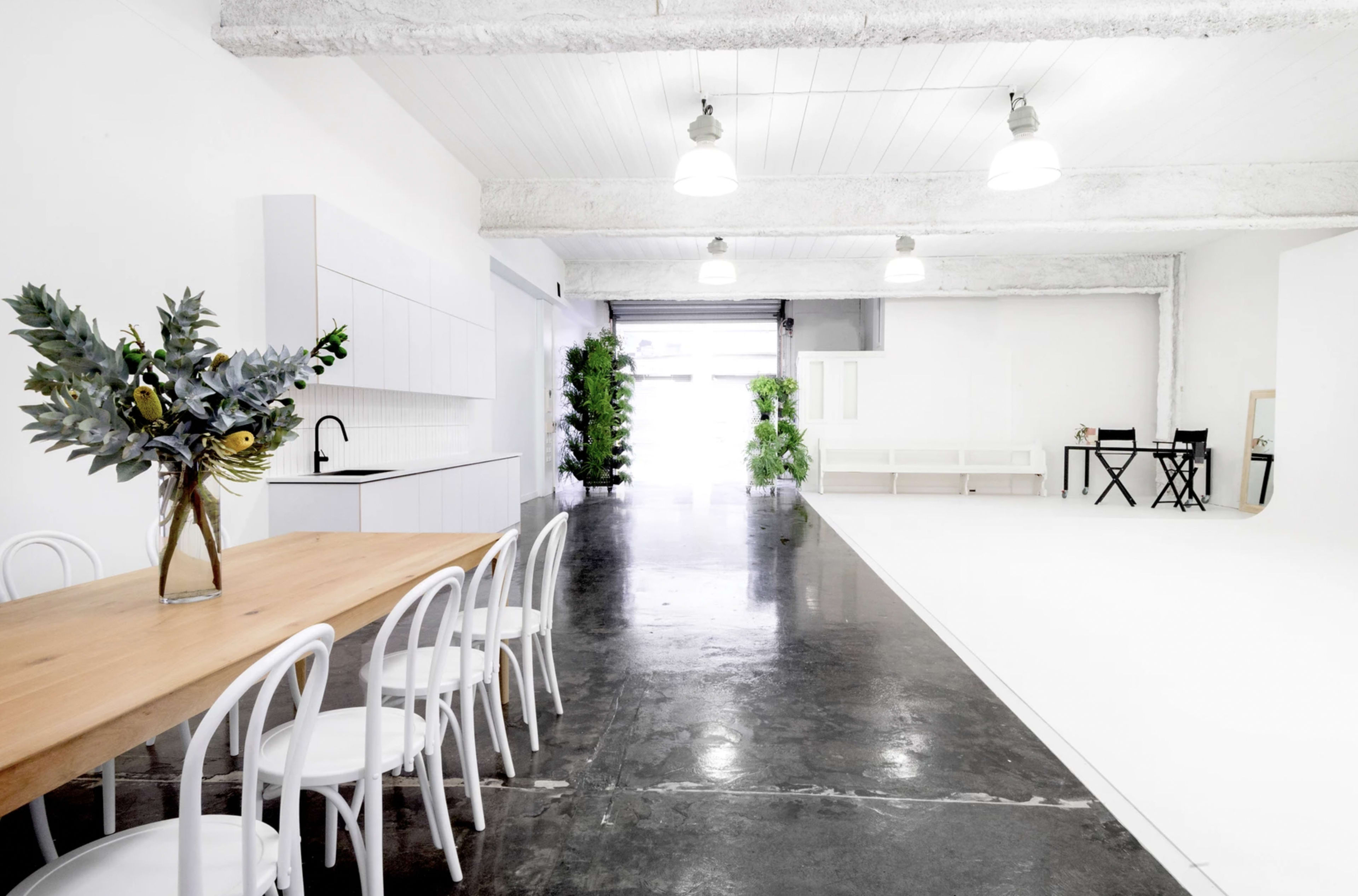 A spacious, modern interior with a long wooden dining table and white chairs, a kitchen area, greenery, and a large white backdrop suitable for photography.