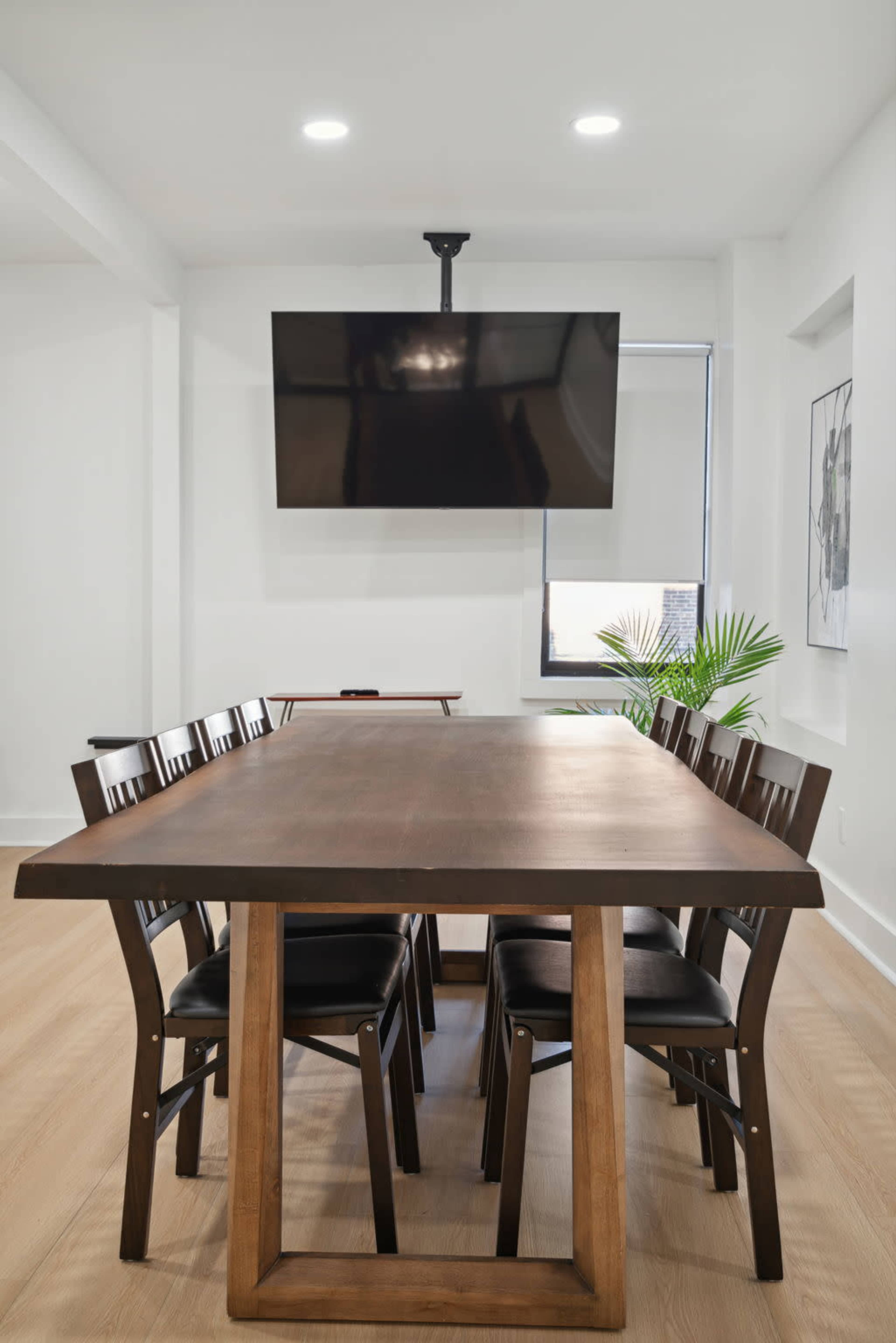 A large wooden dining table surrounded by eight black chairs is positioned in a bright room, with a wall-mounted TV above it and a plant visible in the corner.
