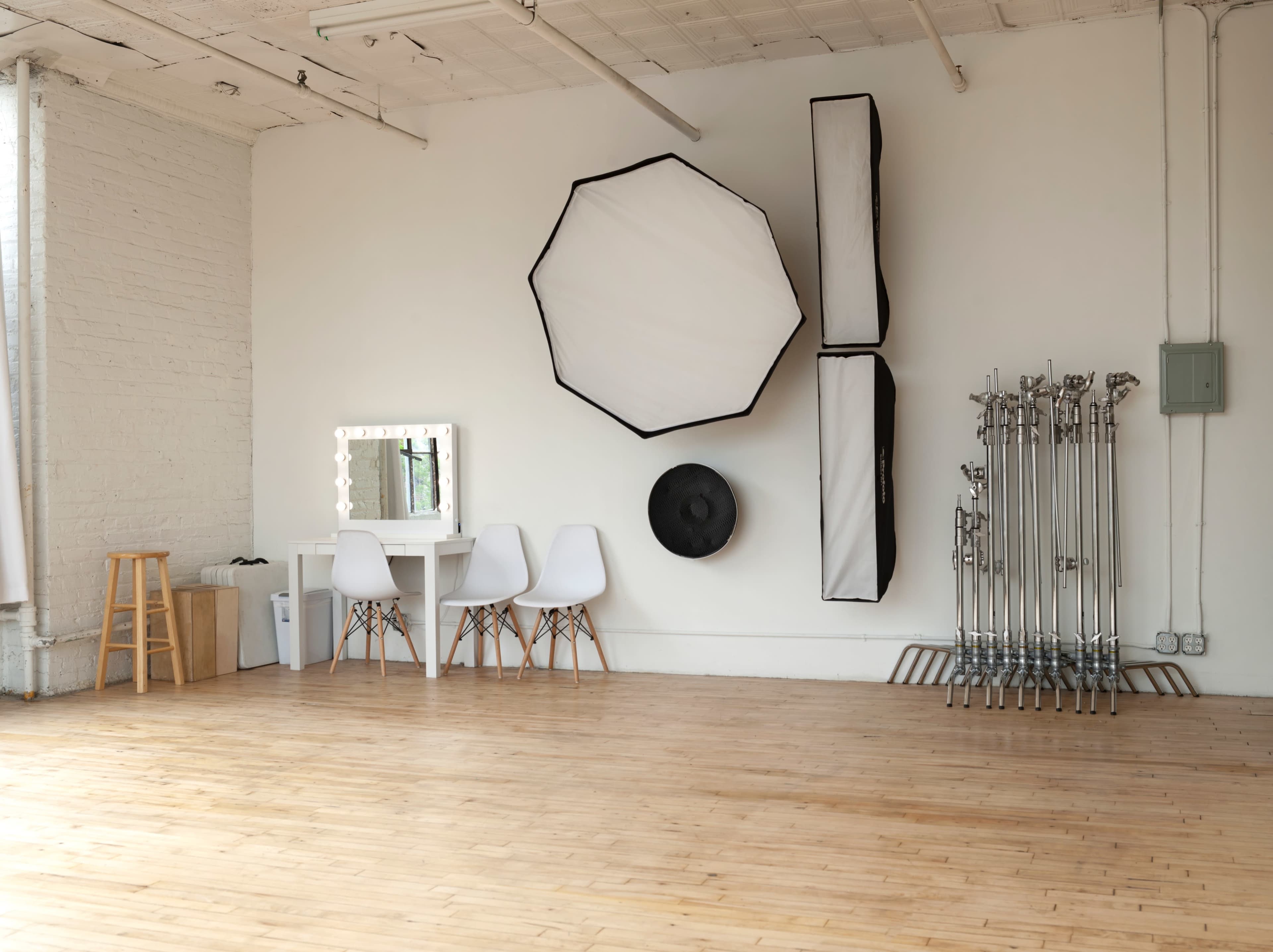 The room features a large wooden floor with a makeup vanity, white chairs, and various photography lighting equipment mounted on the wall.