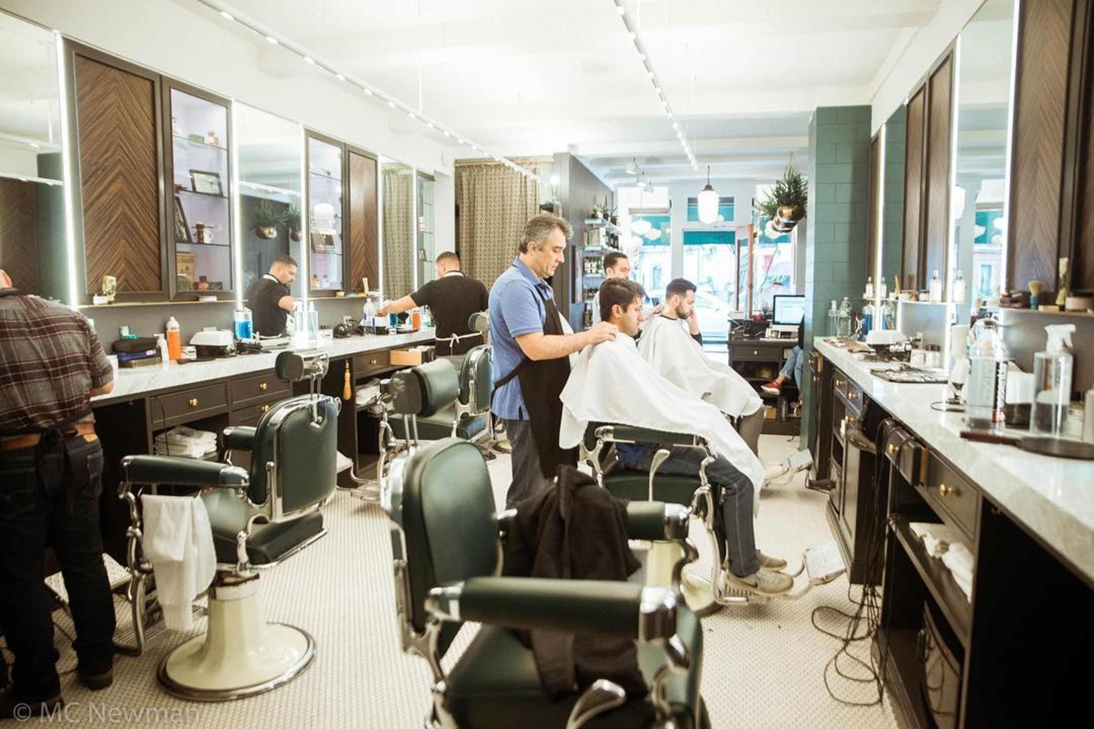 A barber is cutting the hair of a man seated in a grooming chair inside a barbershop with multiple workstations.