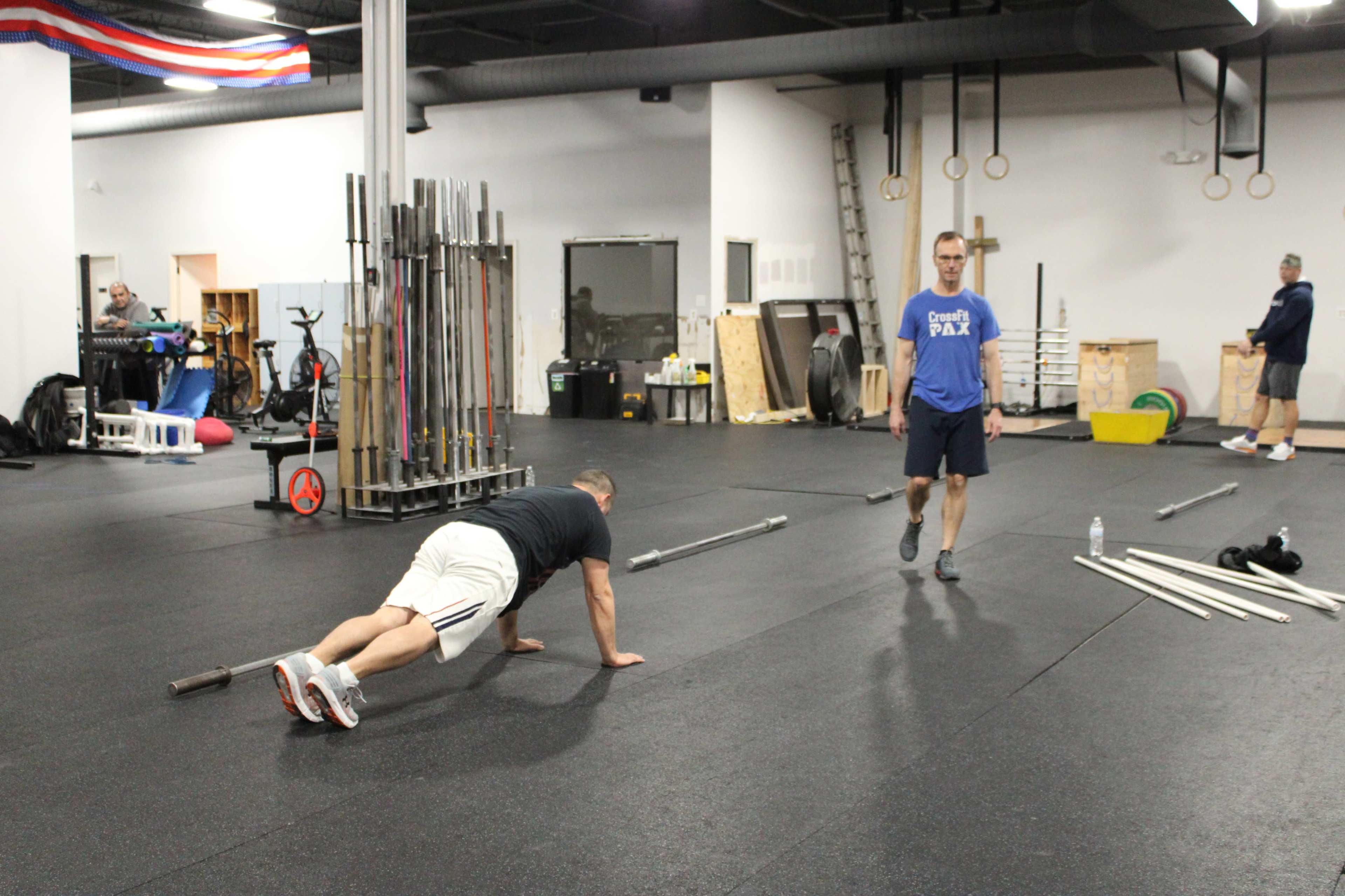 Two men are exercising in a gym, with one doing push-ups on the floor and the other walking nearby.