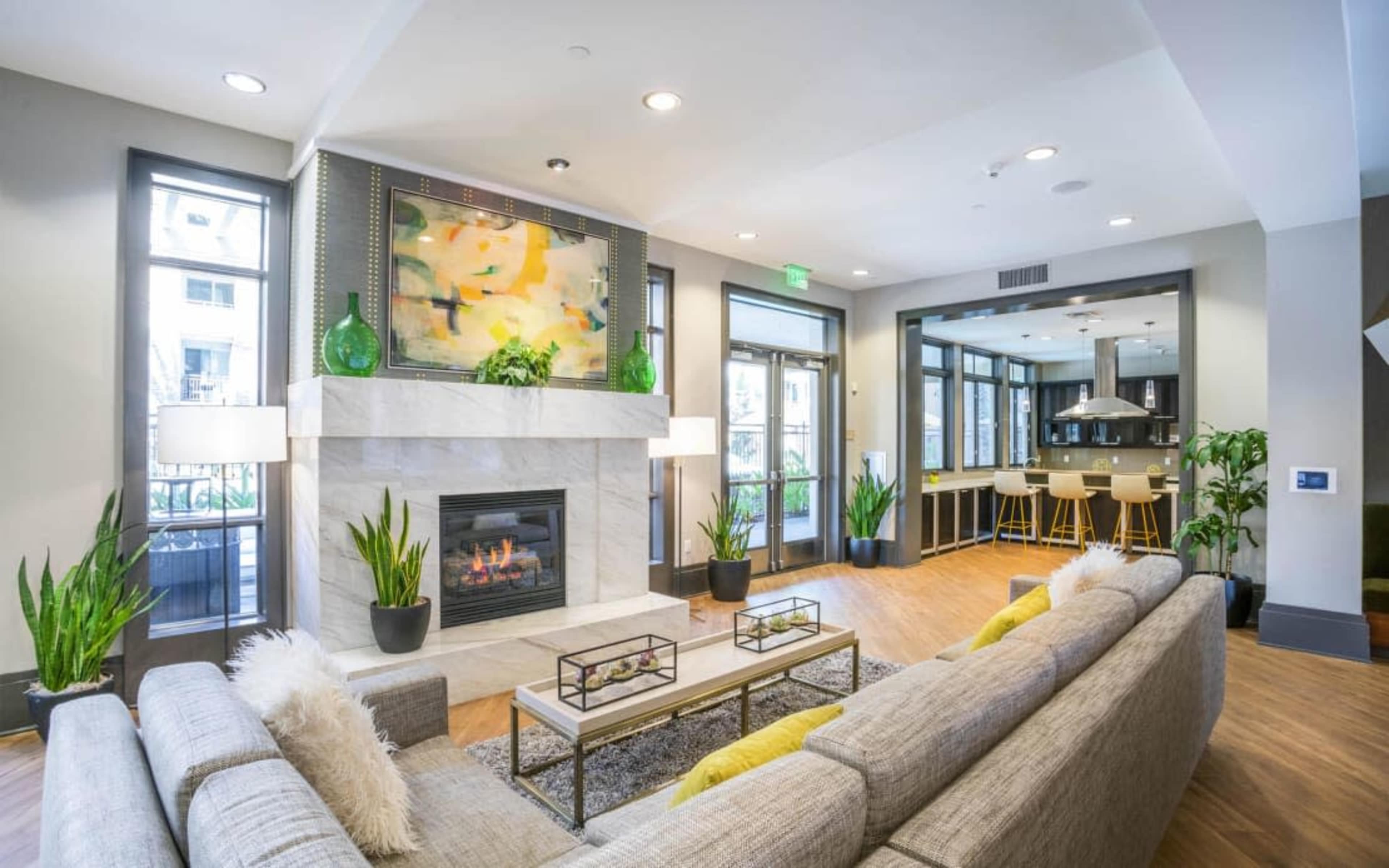 A modern living room features a fireplace, a large gray sectional sofa, and a vibrant painting above the mantel, complemented by plants and a kitchen area in the background.
