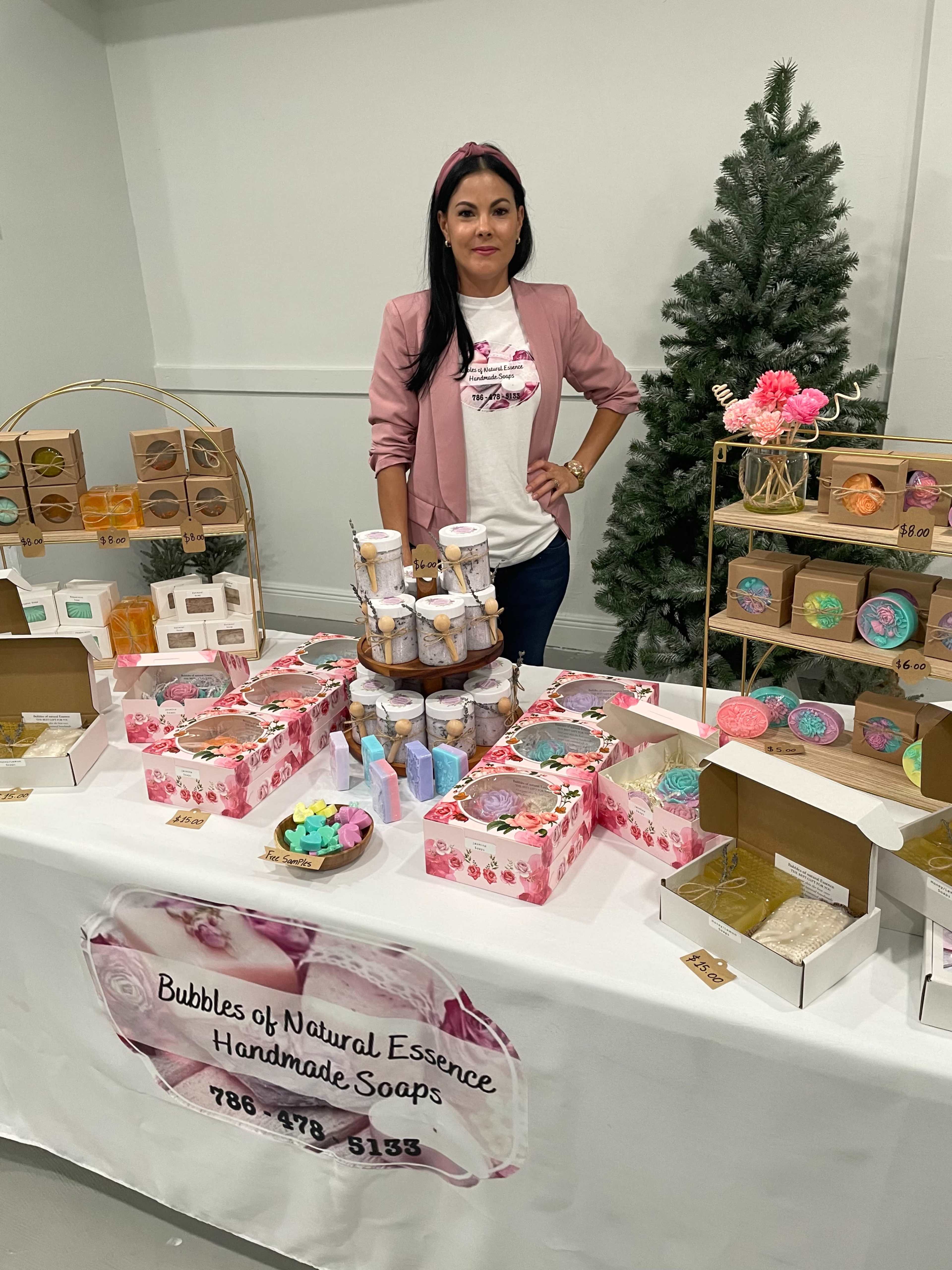 A woman stands behind a table displaying an array of handmade soaps and bath products at a craft fair.