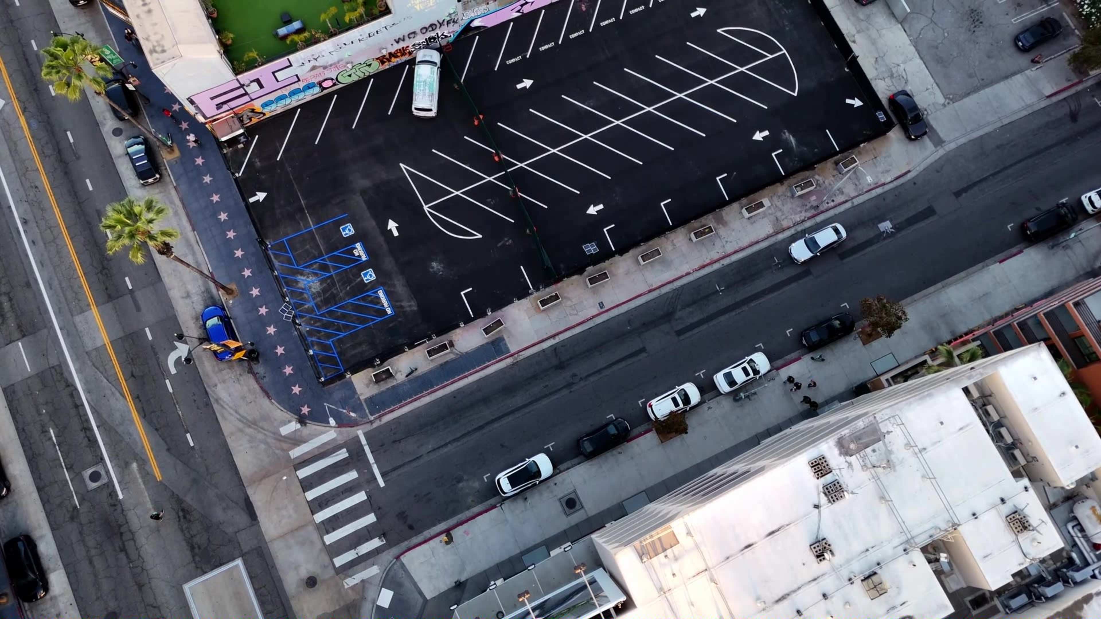 The image shows an aerial view of a parking lot with marked parking spaces adjacent to a street lined with parked cars.