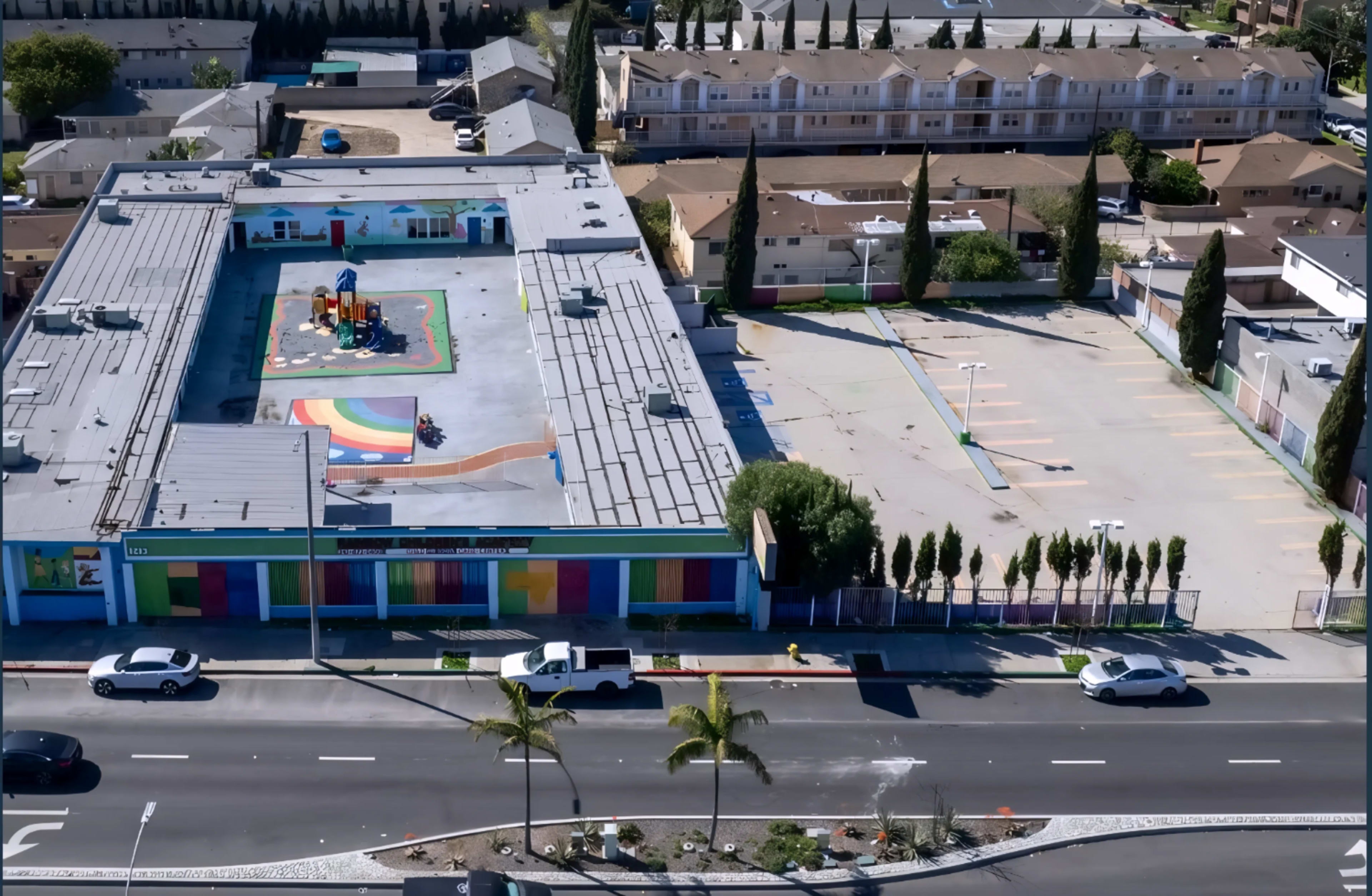 The image shows an aerial view of a colorful building with a playground featuring a rainbow design, surrounded by a parking area and several trees.