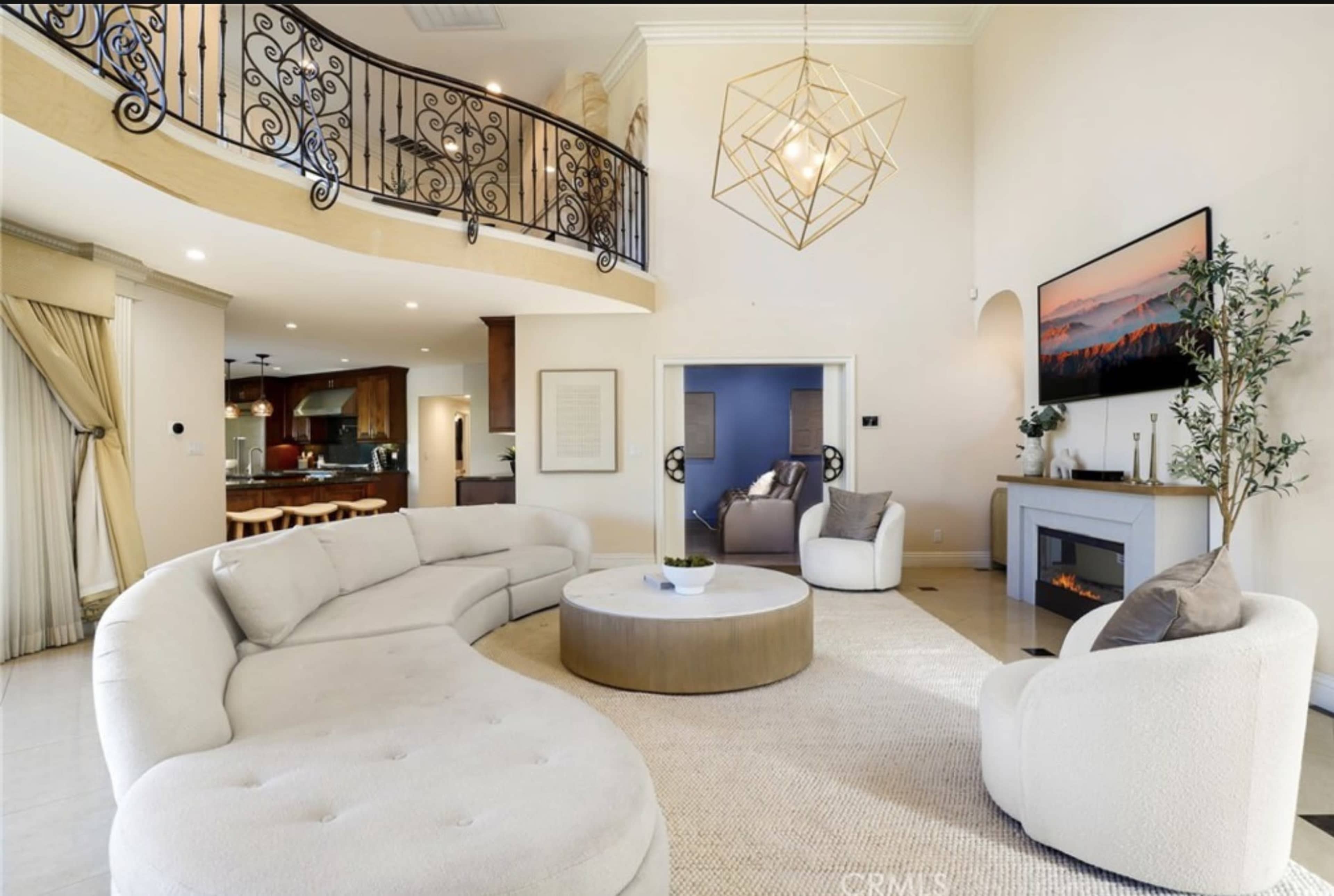 The image features a modern living room with a curved sectional sofa, a round coffee table, and a fireplace, accompanied by a balcony and an open kitchen area in the background.