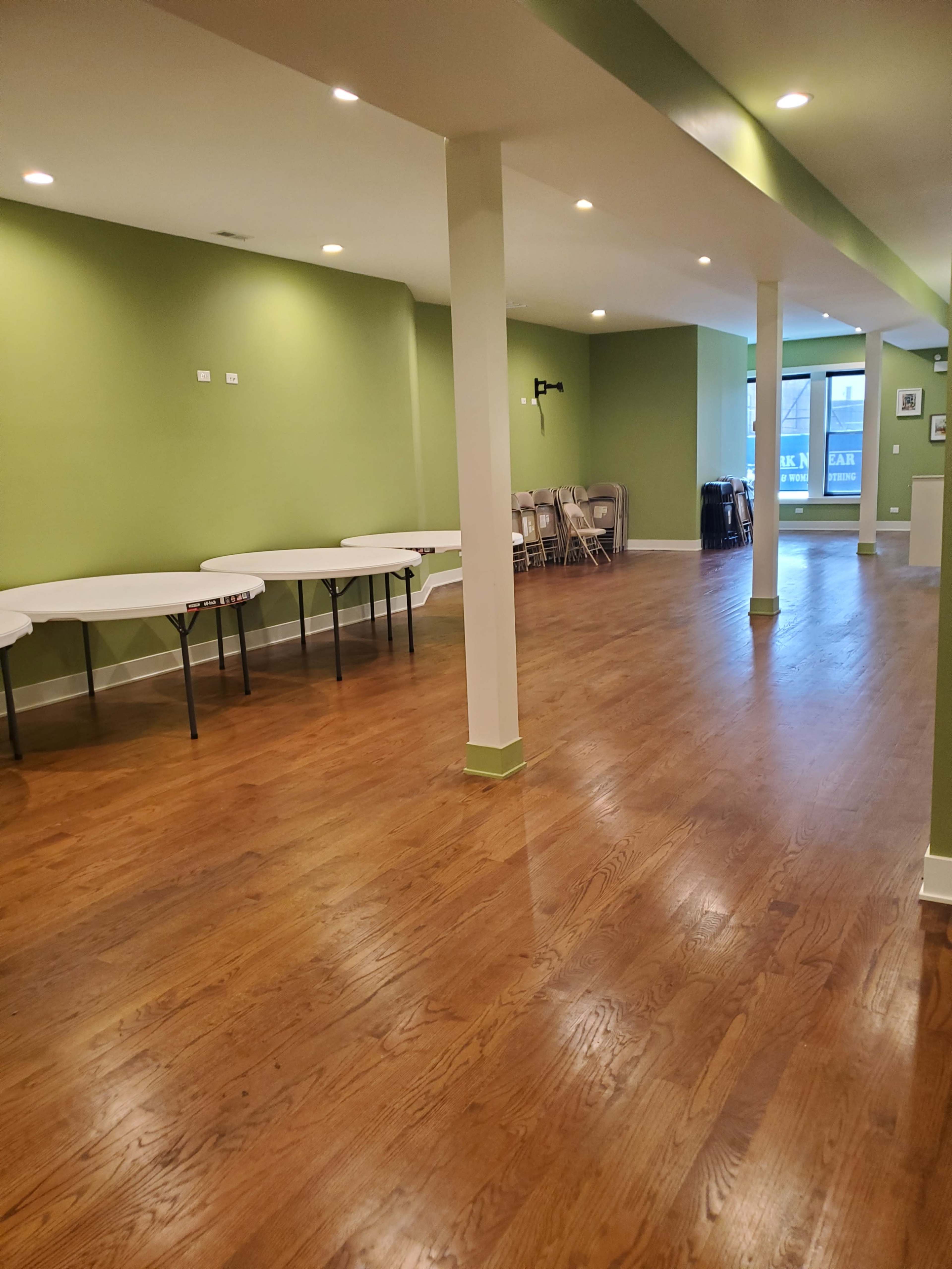 The image shows a spacious, green-walled room with wooden flooring, featuring several tables along one side and chairs stacked in the background.