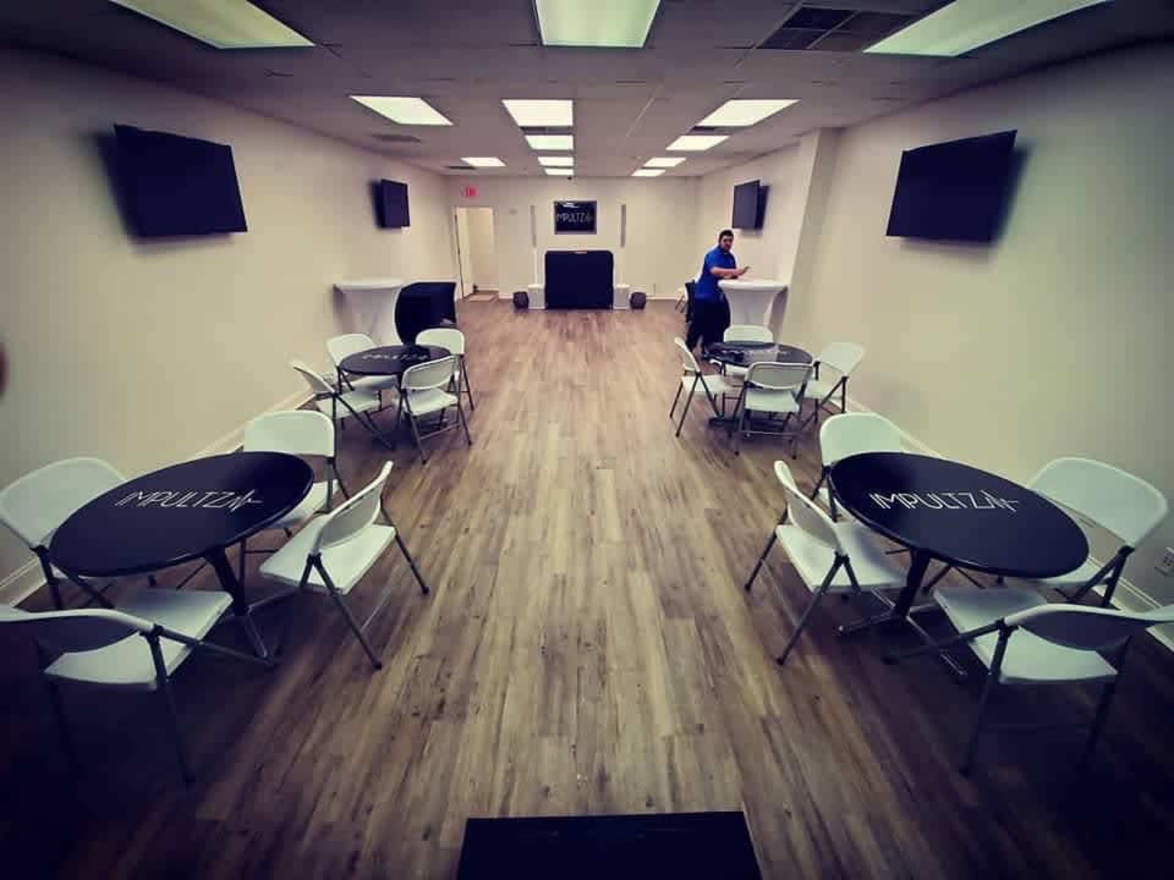 A spacious, well-lit event room with several round black tables and white chairs arranged throughout, along with two wall-mounted televisions and a small stage at the back.