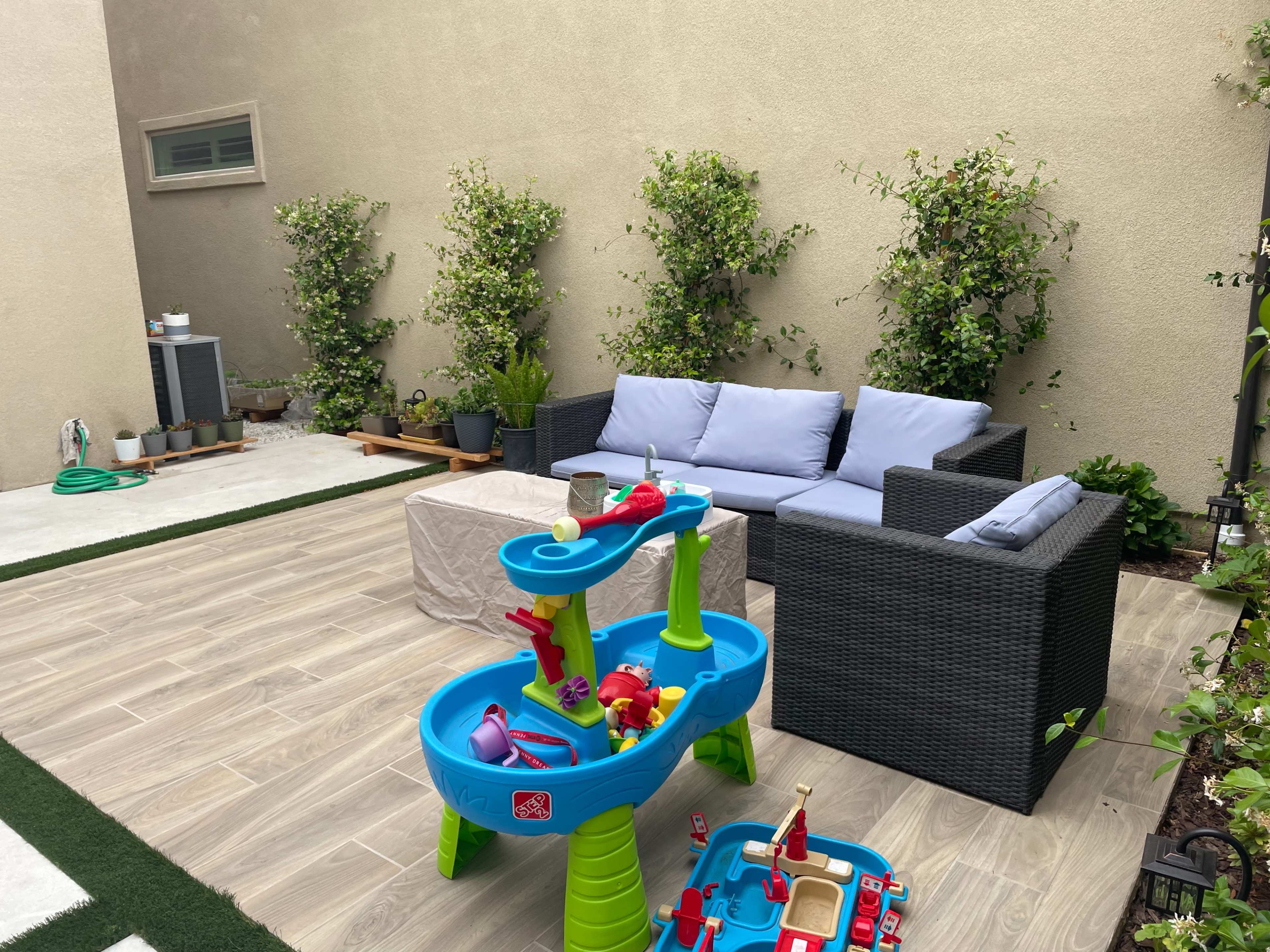 A small outdoor patio with a couch, a coffee table, and a children's play table surrounded by potted plants and greenery.