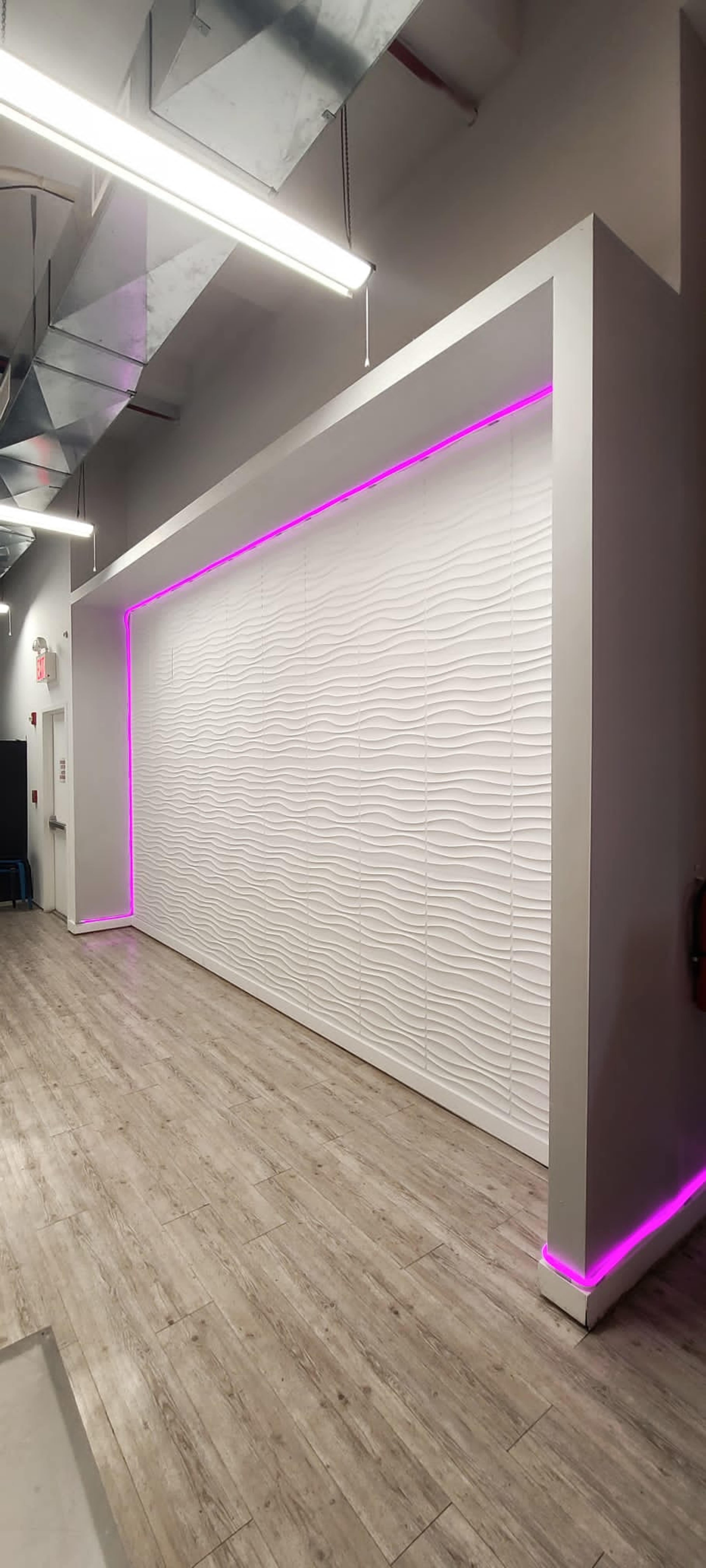 A modern wall with a textured white surface illuminated by pink ambient lighting along its bottom edge, set in a room with a wooden floor.