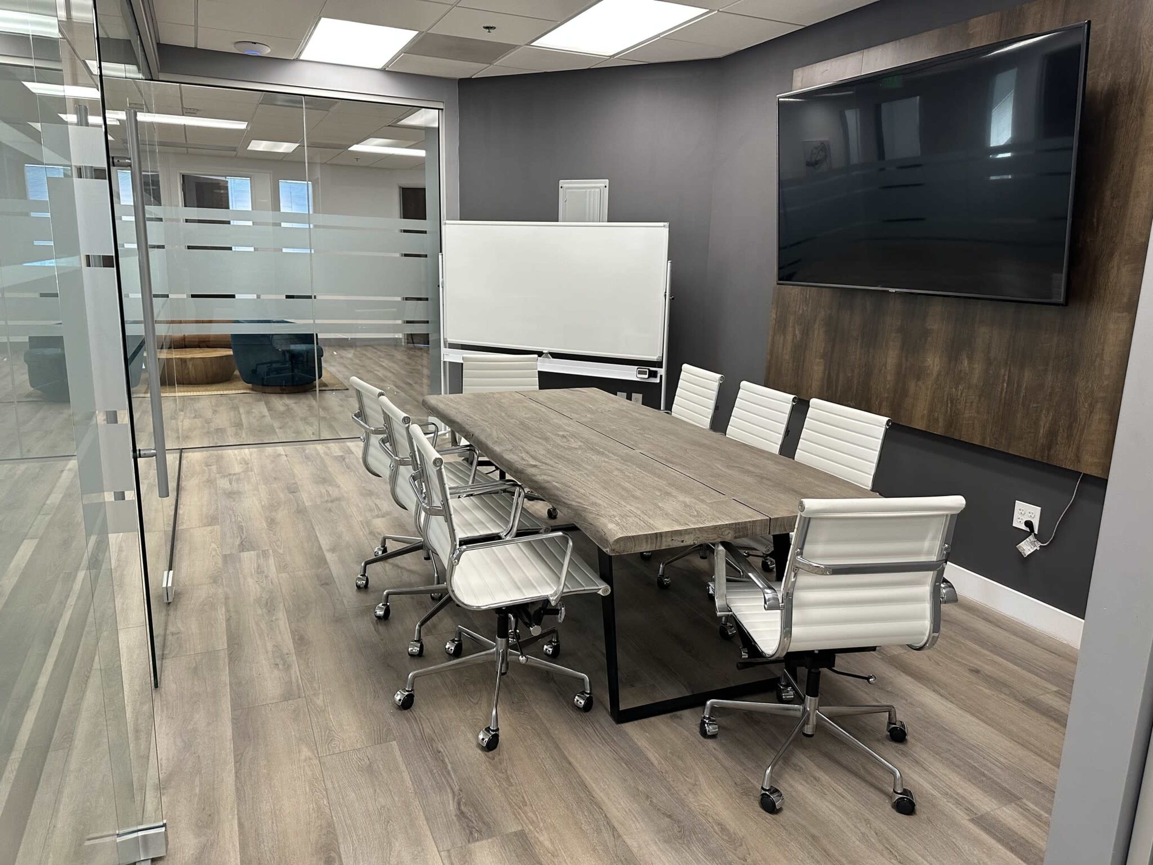 The image shows a modern conference room featuring a large wooden table surrounded by white office chairs, with a wall-mounted TV and a whiteboard.