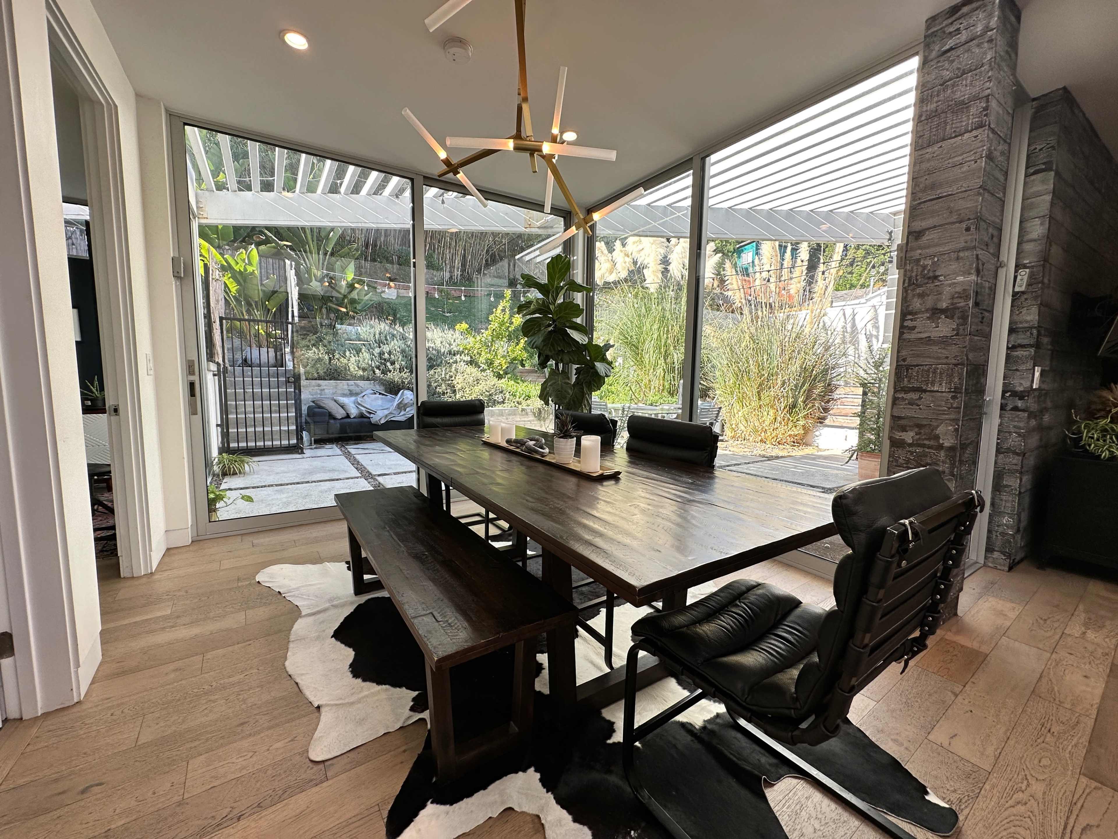 A modern dining area features a large wooden table with black chairs and a bench, surrounded by glass walls that overlook a landscaped outdoor space.